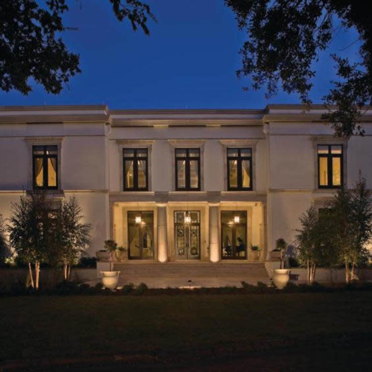 A large, modern two-story house illuminated at night, with symmetrical windows and landscaped greenery in front.