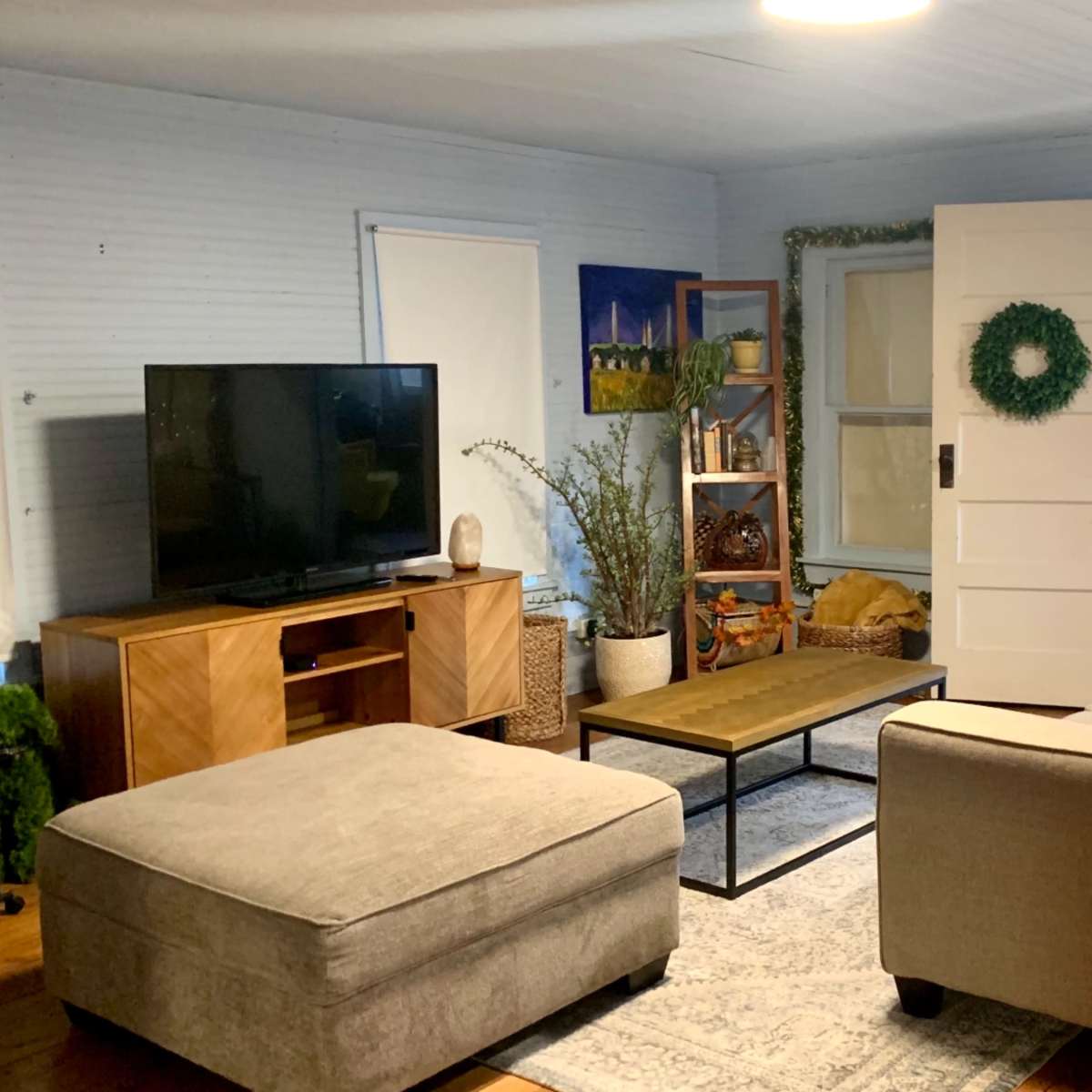 A living room with a television on a wooden console, a beige sofa and ottoman, a coffee table, and decorative greenery along the walls and furniture.