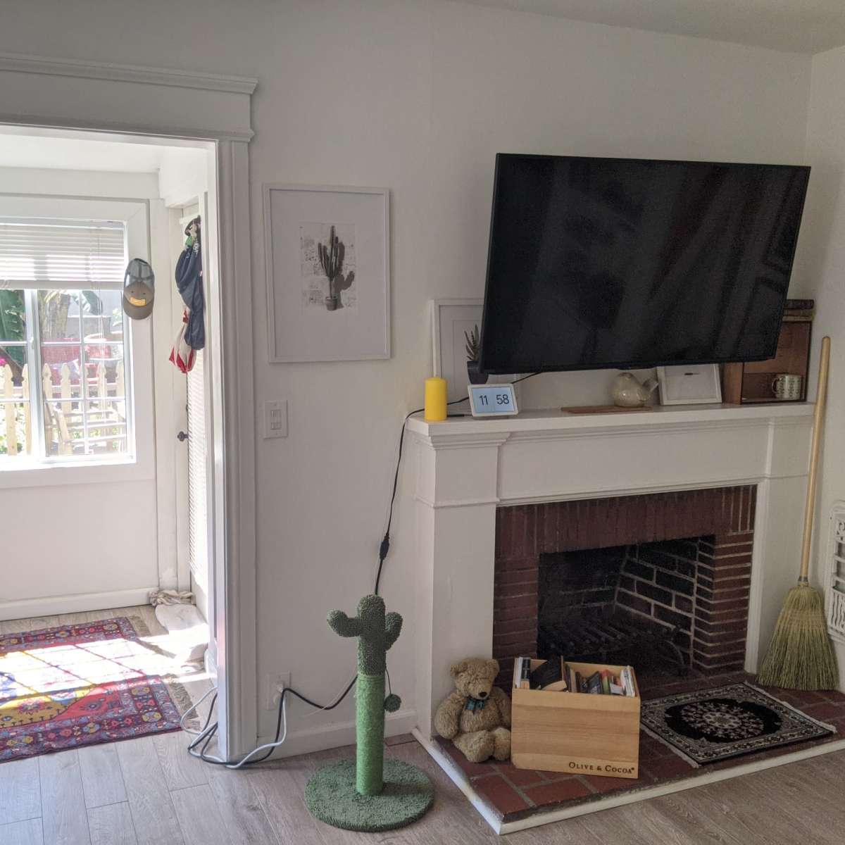 A living room featuring a large TV mounted above a fireplace, a decorative cactus, a wooden box with toys, and an adjacent sunlit space with a window.