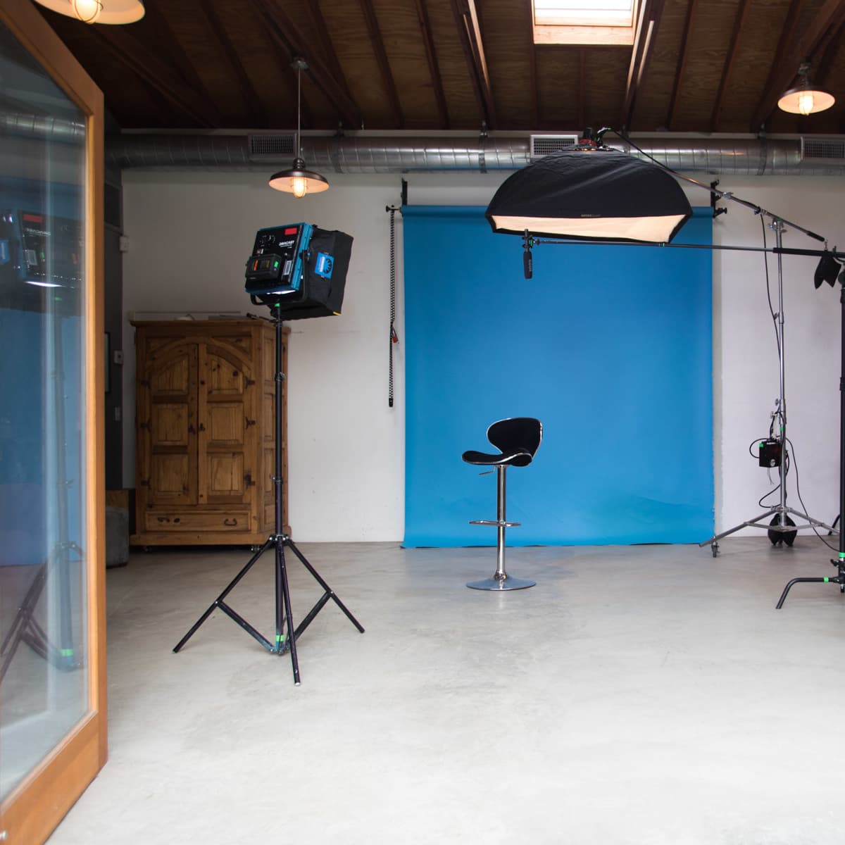 A photography studio with a blue backdrop, lighting equipment, and a single black stool positioned center stage.
