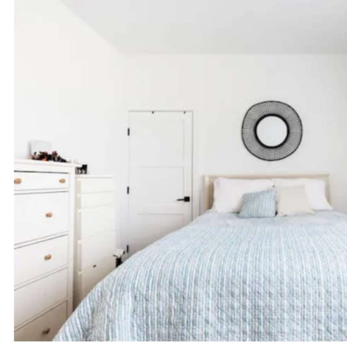 A minimalist bedroom with a bed covered in a light blue quilt, a round mirror on the wall, and a small shelf above a dresser.