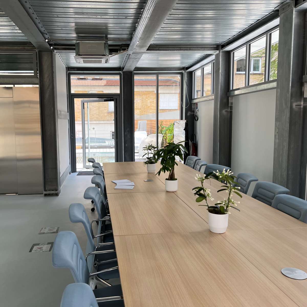 A large conference room features a long wooden table surrounded by light blue chairs and plants, with glass walls and an elevator visible in the background.