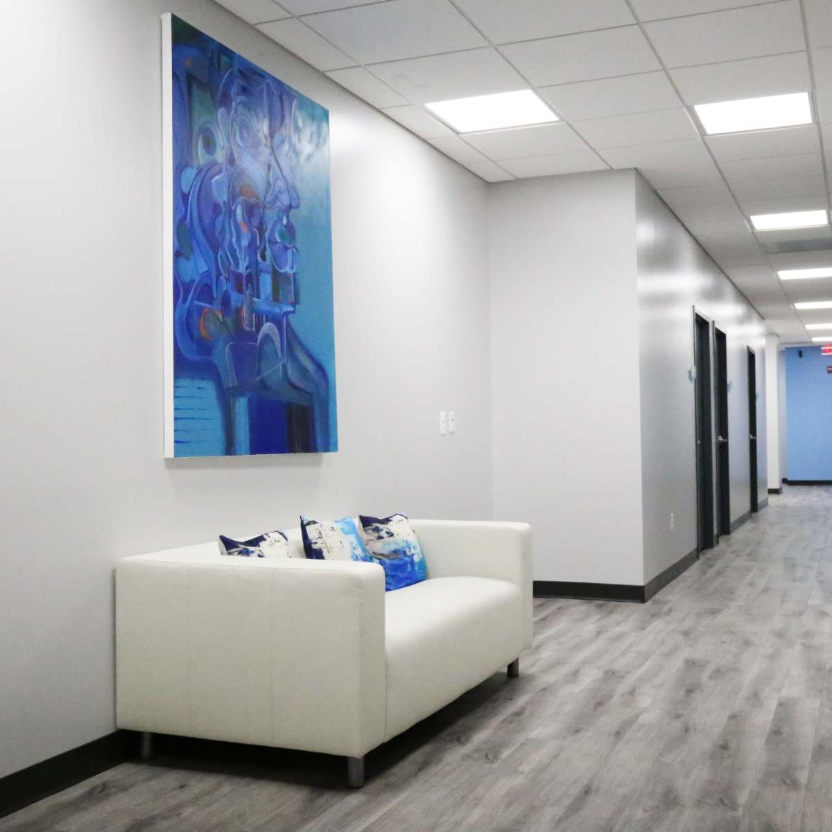 A white sofa with decorative pillows sits against a wall in a long, well-lit hallway featuring several doors and a large piece of abstract art.