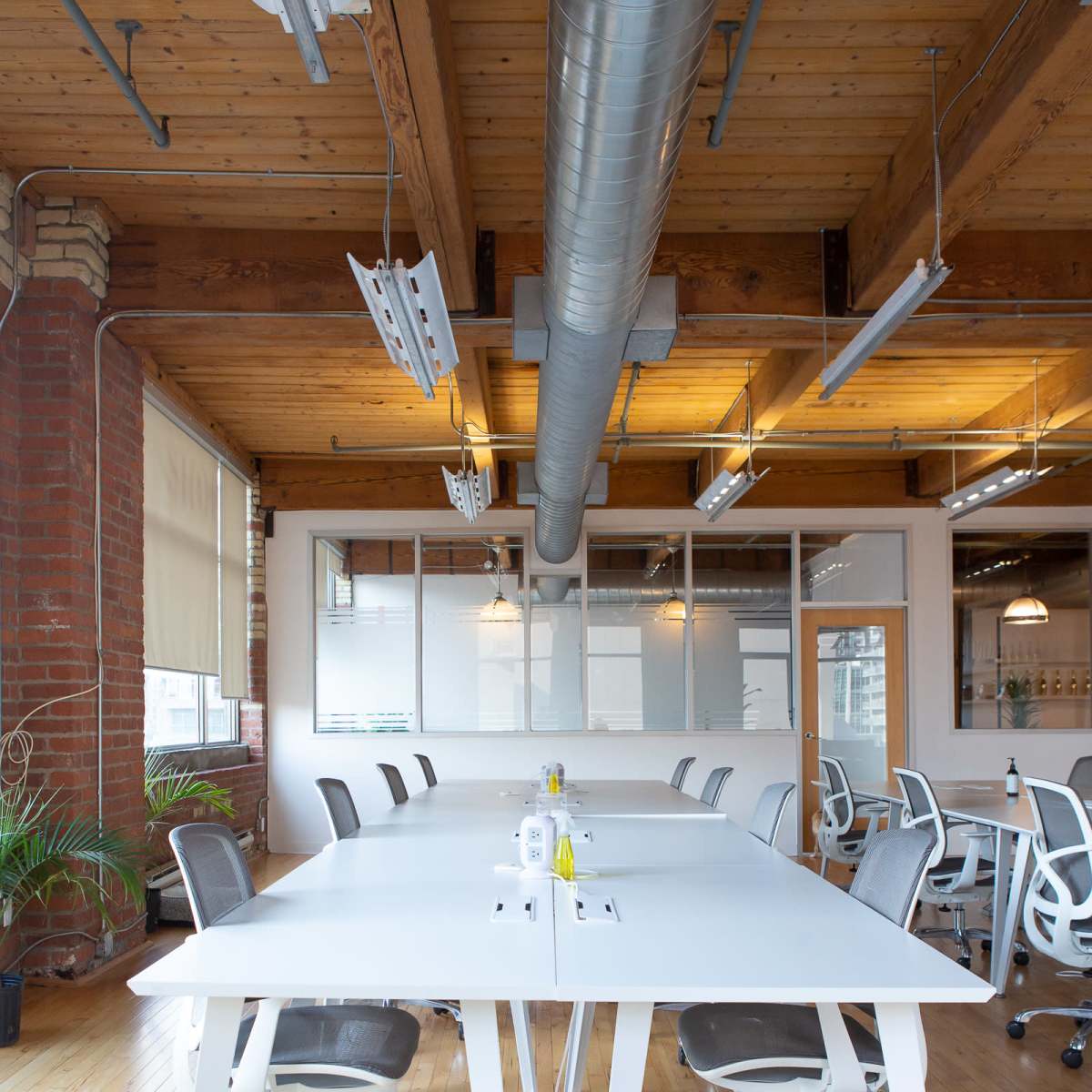 A modern office space features a large white table surrounded by ergonomic chairs, with wooden floors and exposed brick walls.