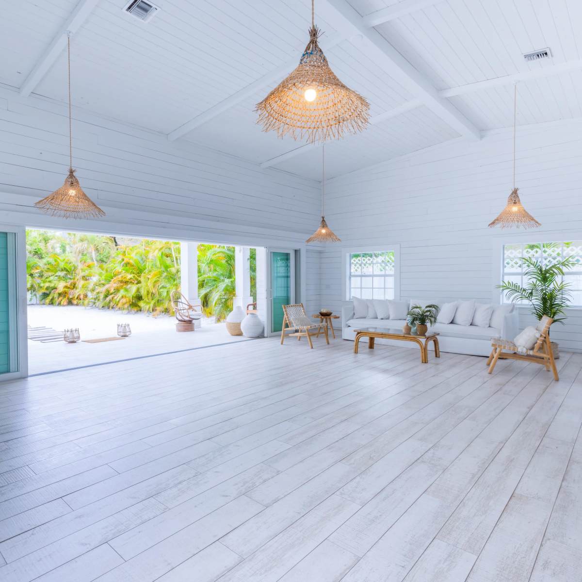 A spacious, brightly lit interior features white wooden walls, a seating area with a bamboo sofa, and tropical plants visible through large windows.