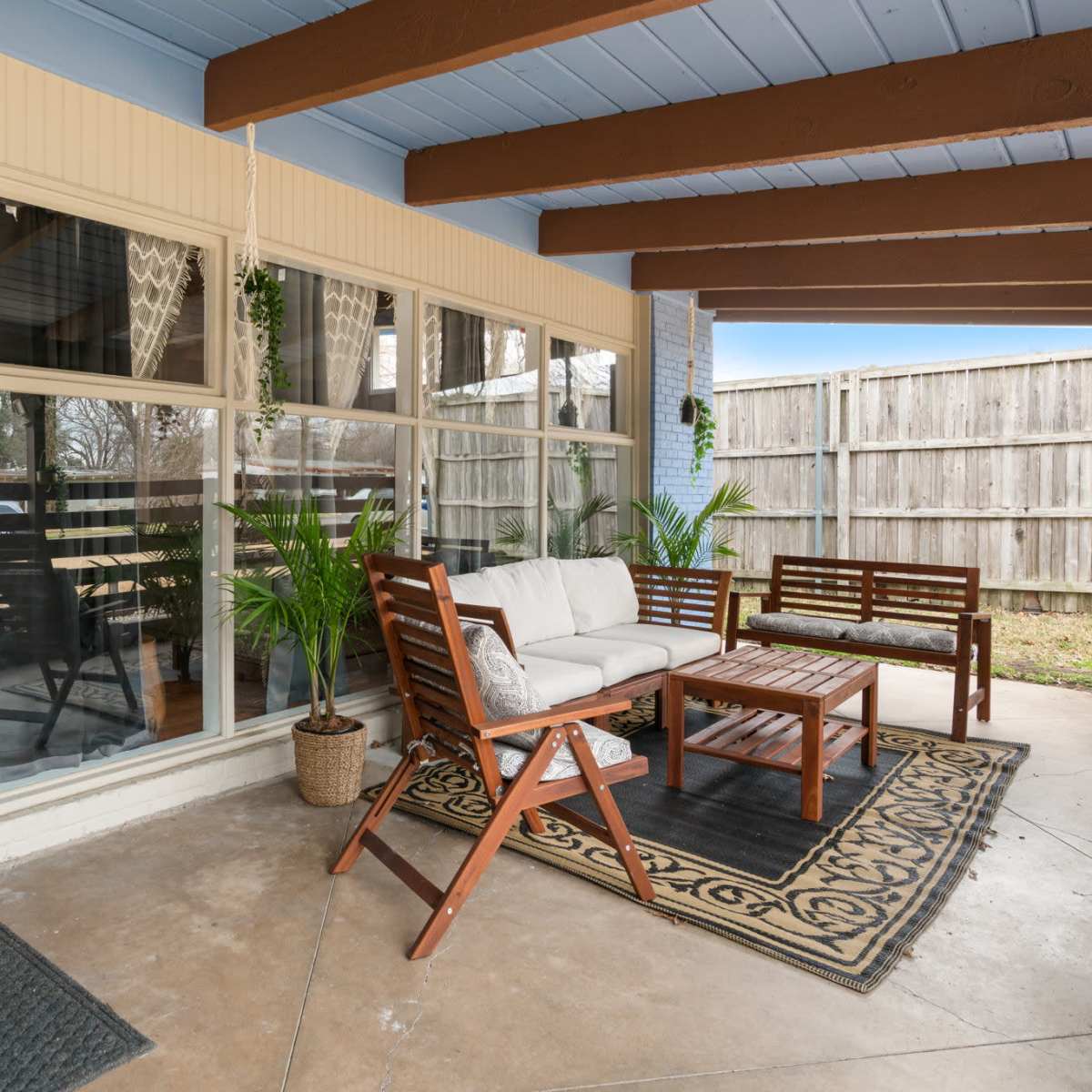 A covered porch with a wooden seating arrangement, a coffee table, and large windows overlooking a backyard enclosed by a wooden fence.