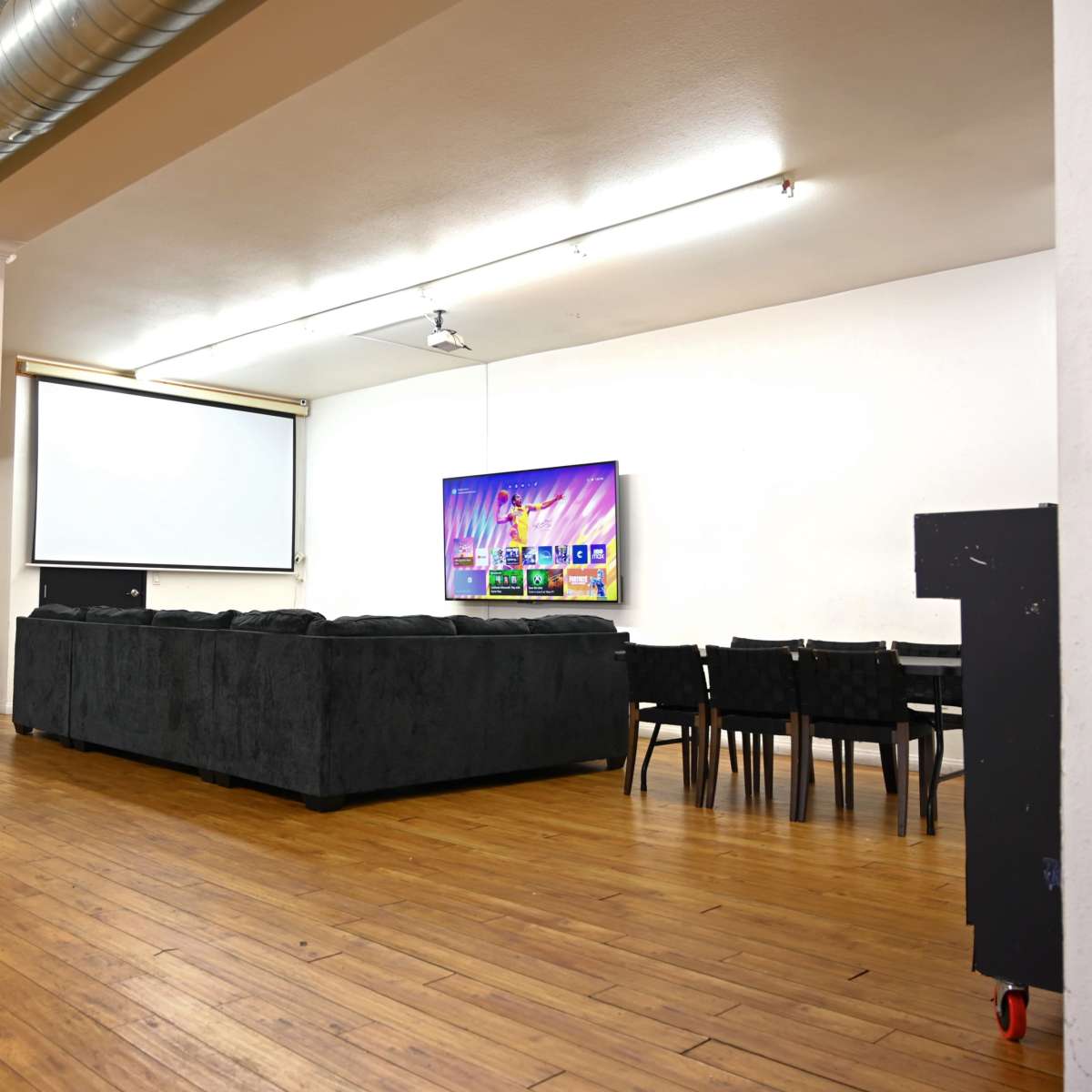 A spacious room with a large sectional sofa facing a wall-mounted television, a projector screen, and a dining area with black chairs.