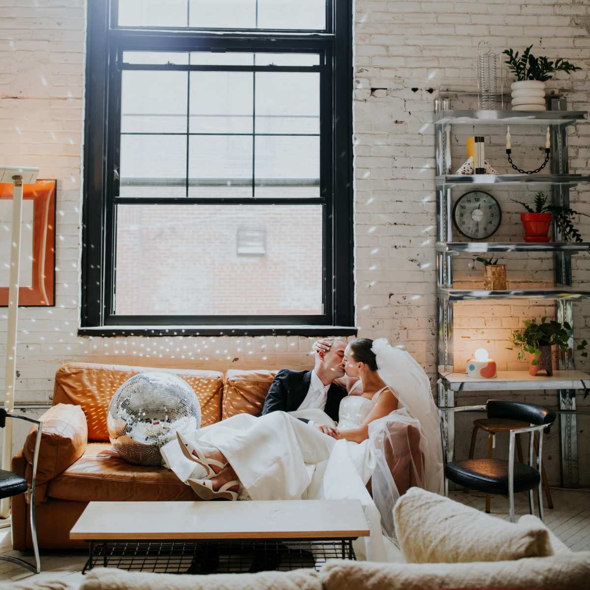 A bride and groom sit together on a couch in a room with large windows and brick walls, surrounded by plants and decorative items.