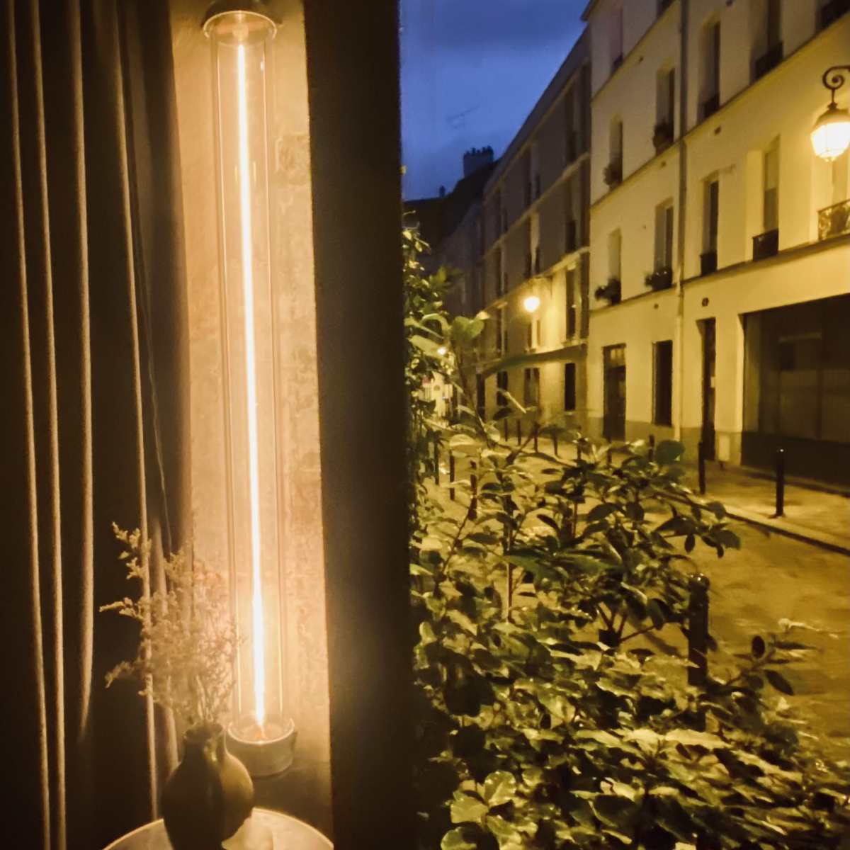 A tall, illuminated lamp stands beside a dark curtain, casting light on a small table with a vase and candles, while outside, a quiet street lined with apartment buildings is visible under evening light.