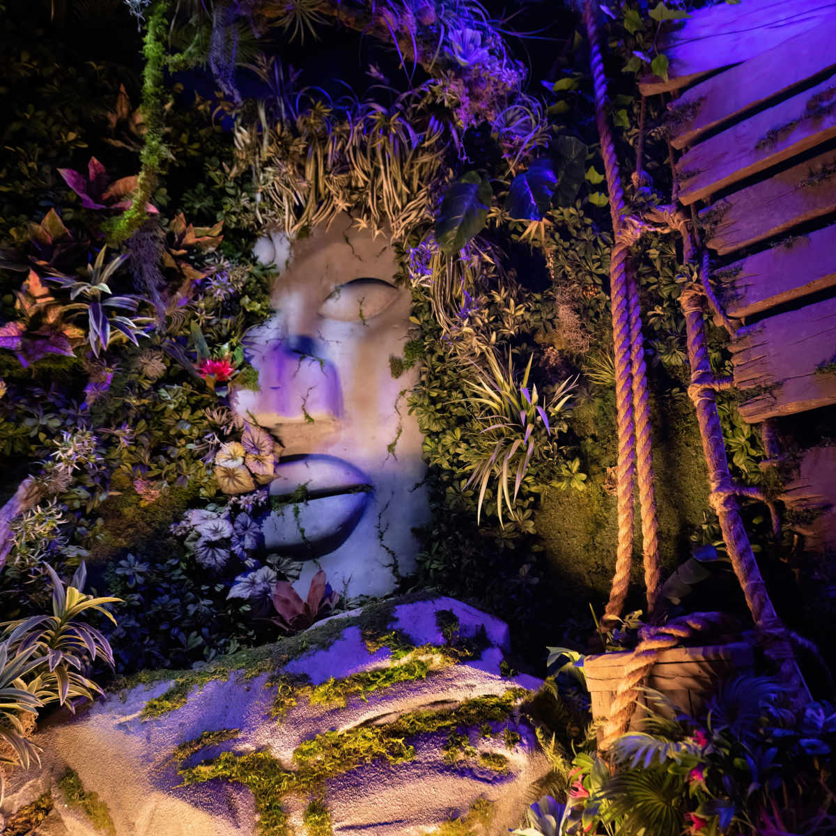 A large, artistic face carved into a rock surface is surrounded by vibrant foliage and illuminated by colored lights, with a wooden structure to the side.