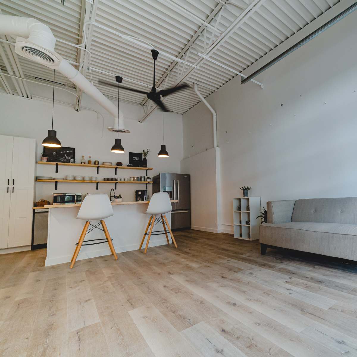 A minimalistic interior space featuring a light-colored sofa, a kitchen island with stools, and large windows along one wall.