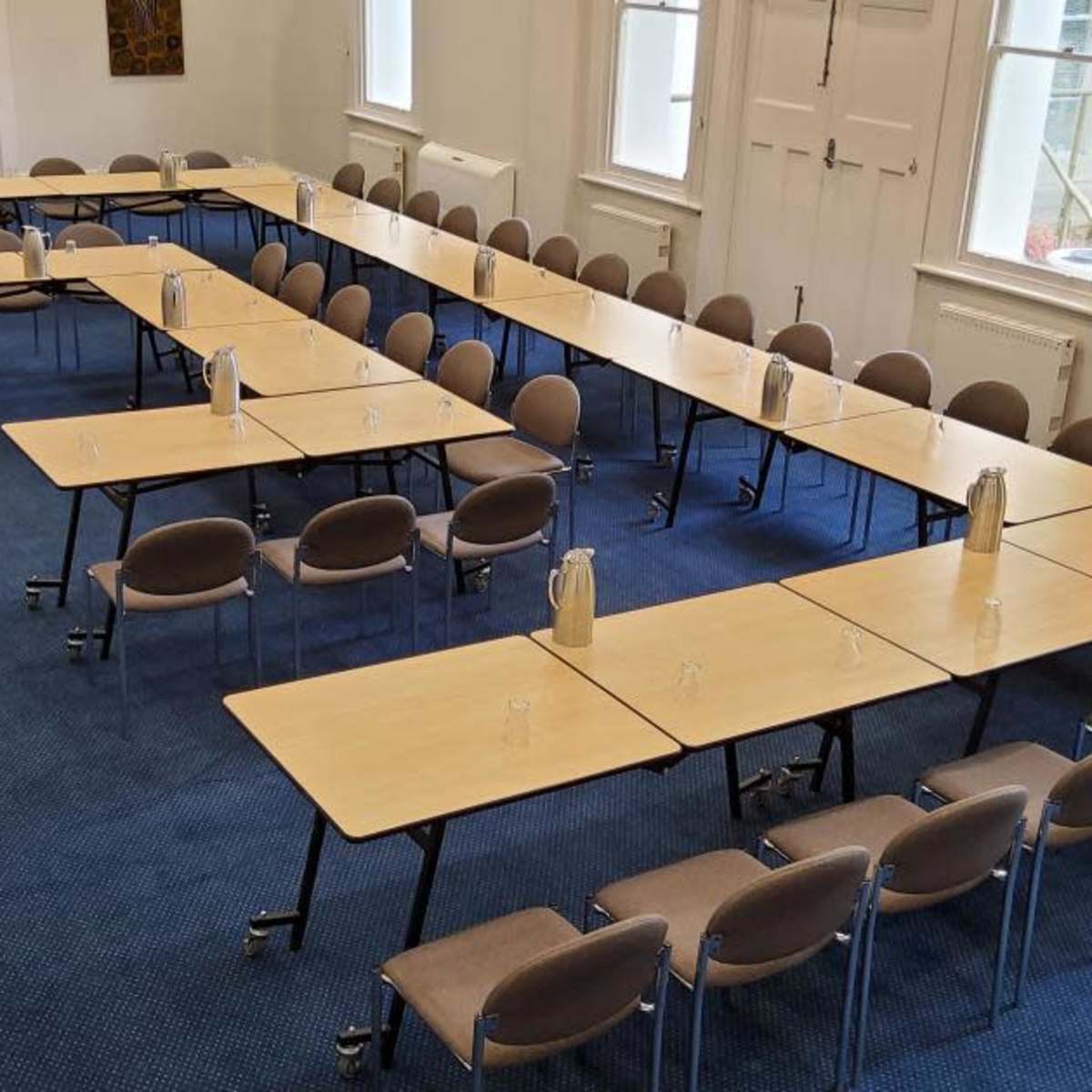 A meeting room arranged with multiple rectangular tables in a U-shape, surrounded by chairs, all on a blue carpeted floor.