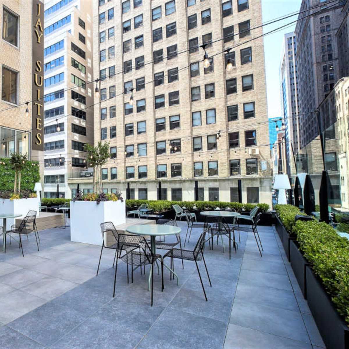 A modern outdoor terrace area with tables and chairs surrounded by greenery and tall buildings.