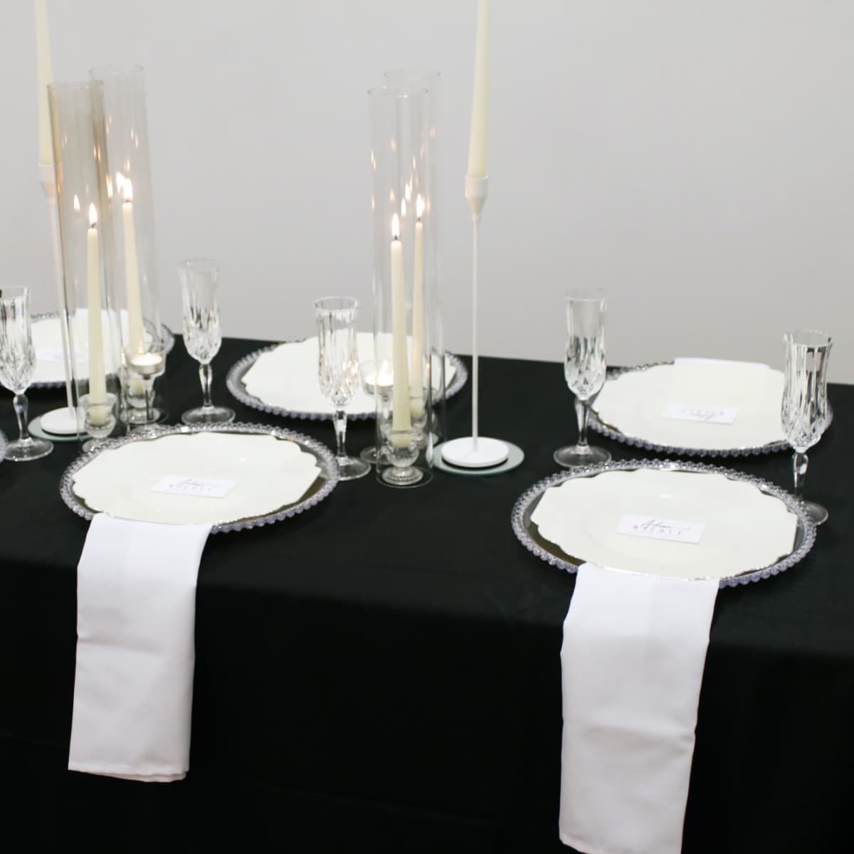 A black tablecloth is set for a formal dinner with white plates, glassware, and tall candles arranged in the center.