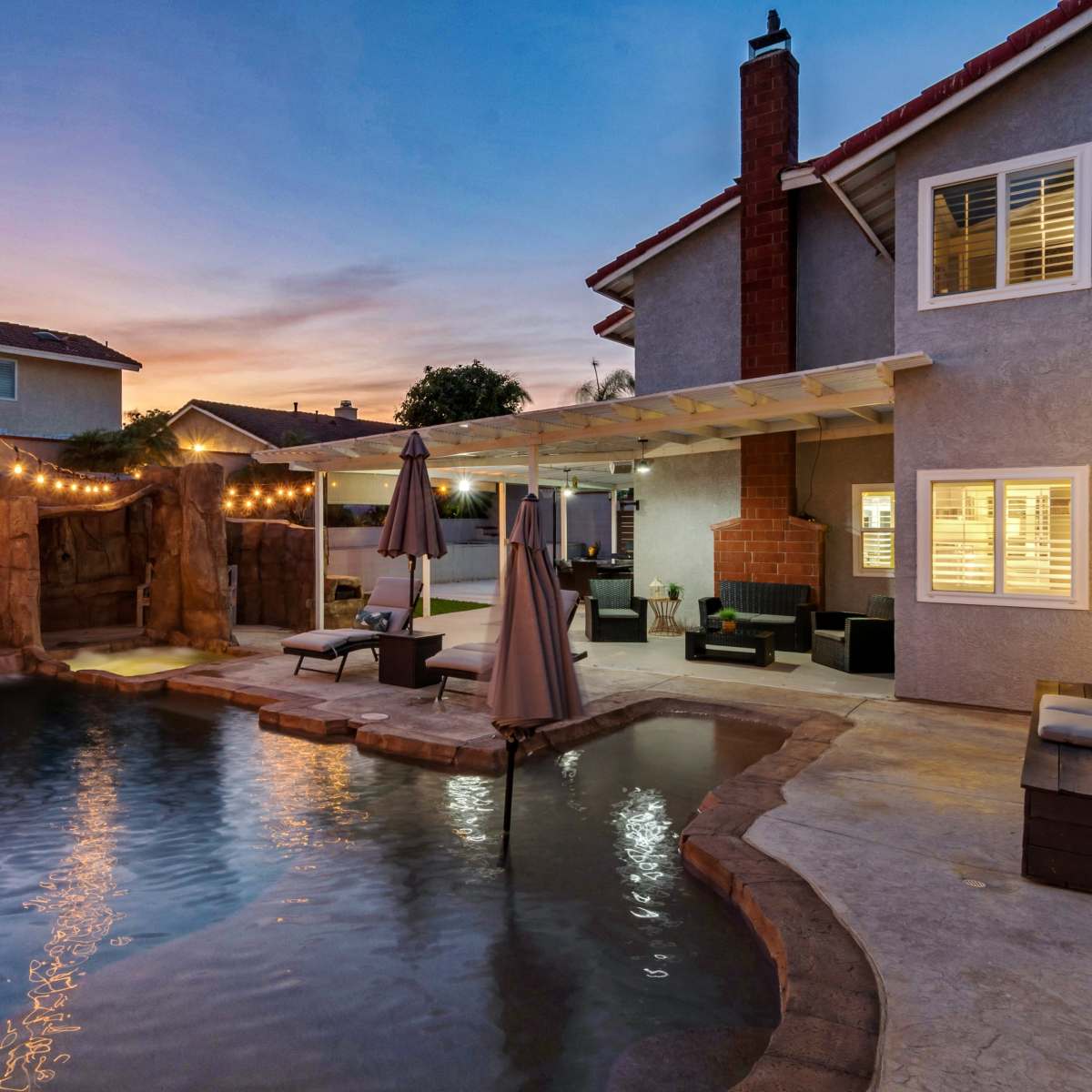 A backyard pool area with rock formations, a waterfall, and string lights illuminating the space during sunset.