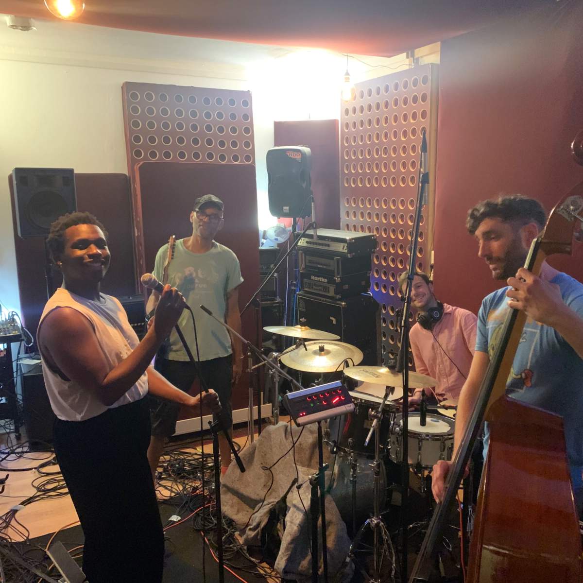 A group of four musicians is playing instruments in a recording studio, surrounded by sound equipment and acoustic panels.