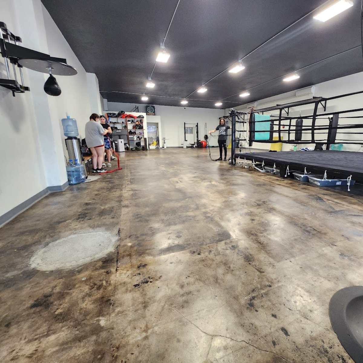 A spacious gym with a boxing ring, training equipment, and individuals standing near a water cooler.