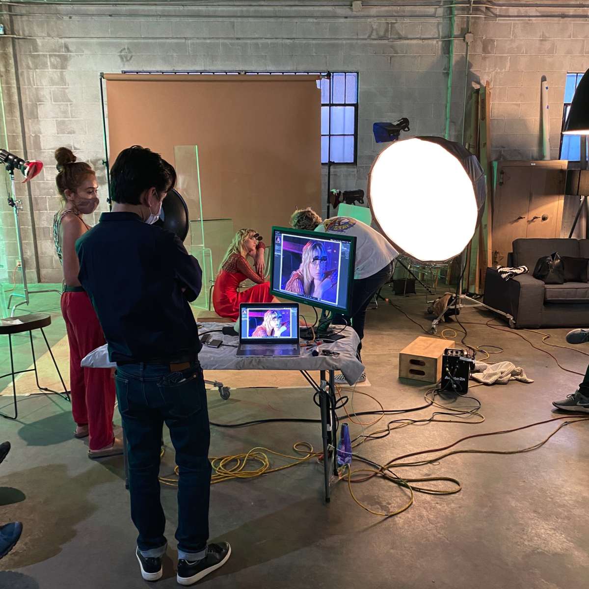A group of individuals is working in a studio, with one person monitoring a video feed on a laptop while others are positioned in front of a large softbox light and a backdrop.