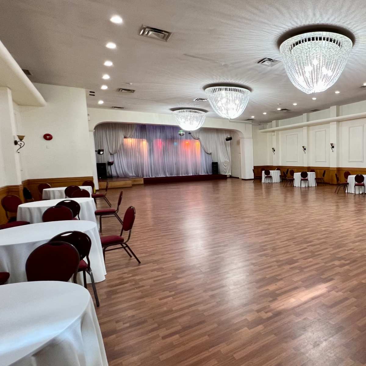 A large, empty event hall features several round tables with chairs and two chandeliers hanging from the ceiling.