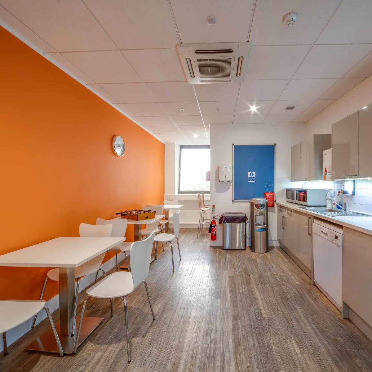 A modern kitchen with an orange accent wall, equipped with a table, chairs, and various appliances including a microwave and dishwasher.