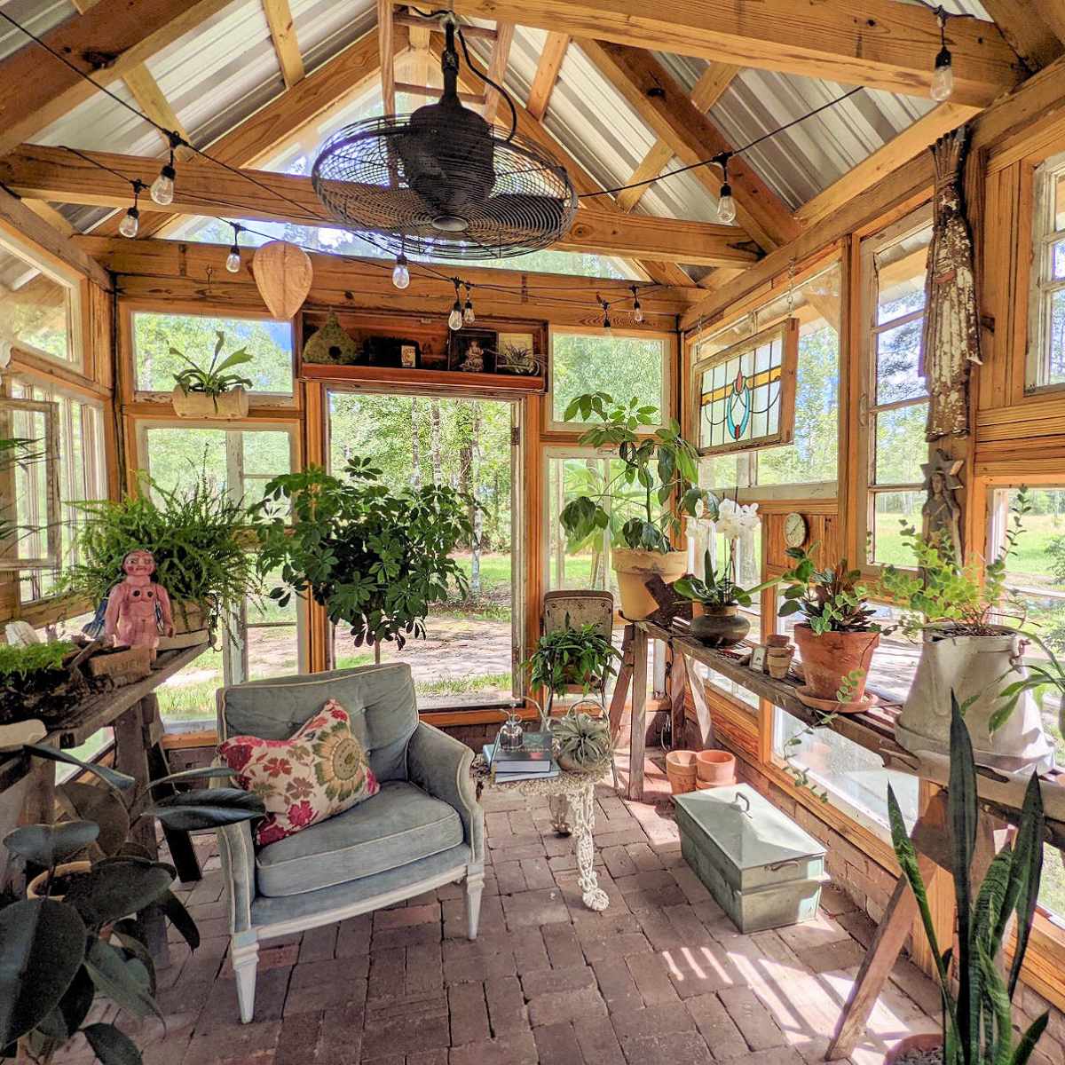 A bright, wooden greenhouse filled with various potted plants and a cozy seating area.