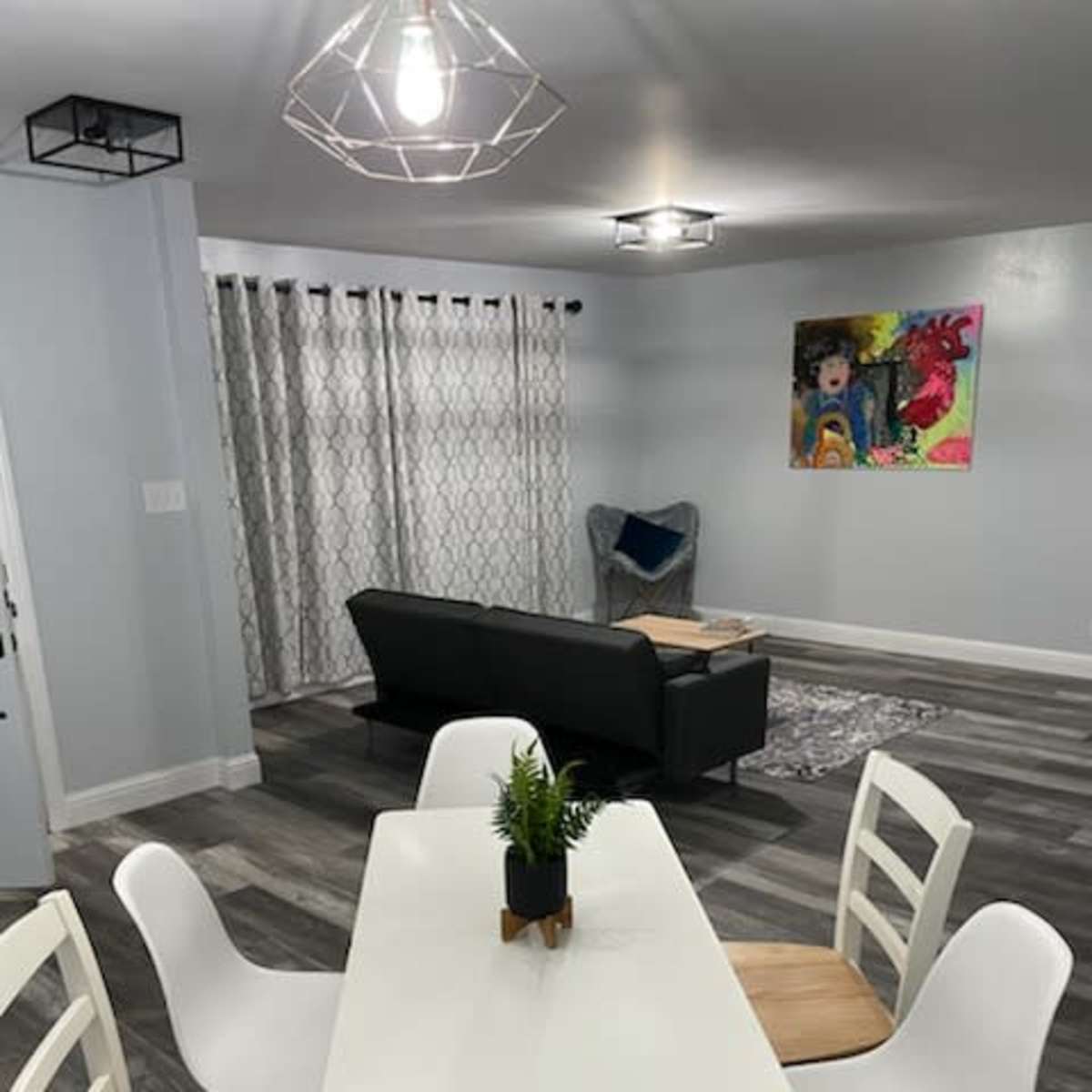 A modern dining area features a white table with wooden accents surrounded by six chairs, with a gray couch and colorful artwork in the background.