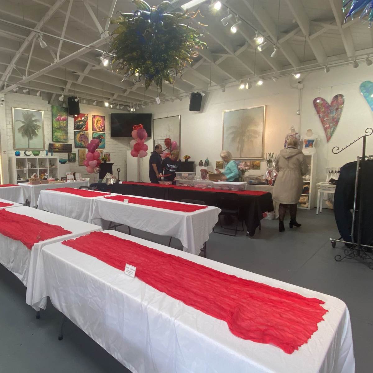 A spacious room with white tables covered in red tablecloths, arranged for an event, while people prepare in the background.
