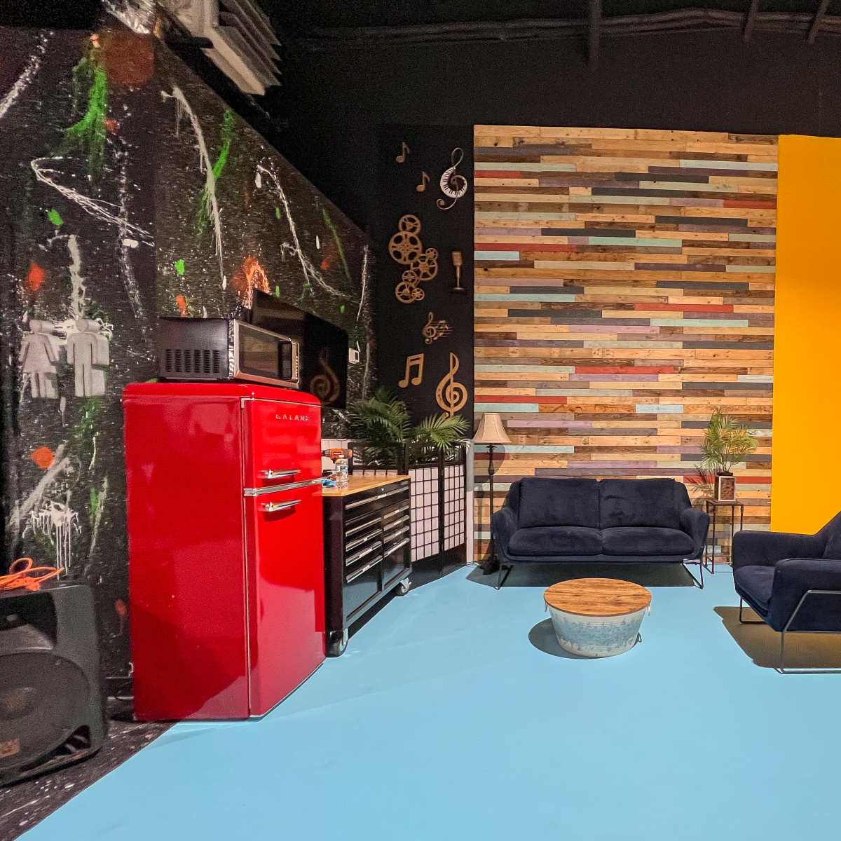 A vibrant studio space featuring a red refrigerator, colorful wooden accent wall, and two dark sofas arranged around a round coffee table on a blue floor.