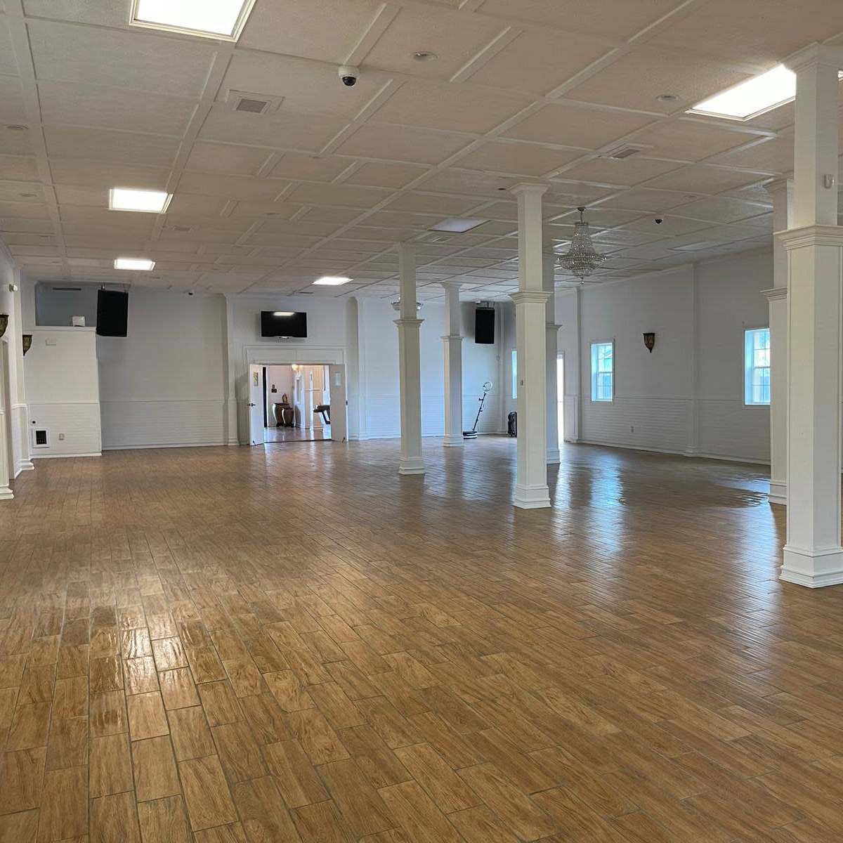An empty, spacious room with wooden flooring, white walls, and tall columns, illuminated by overhead lights.