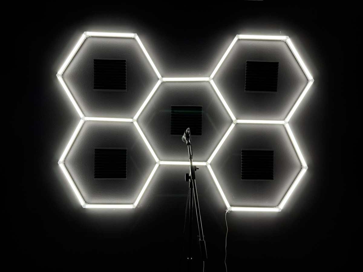 The image shows a dark wall with hexagonal neon lights arranged in a pattern and a microphone on a stand in front of it.