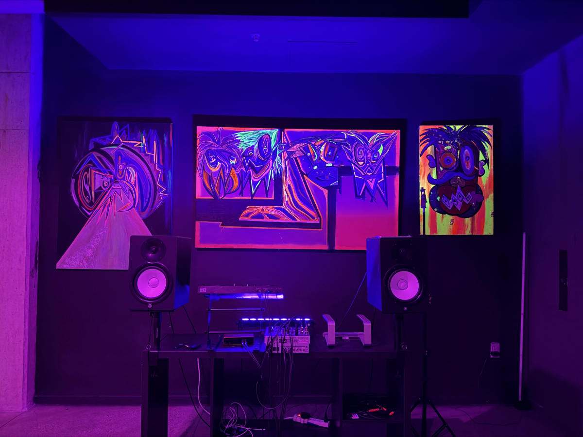 The image shows a dimly lit room featuring four vibrant, abstract paintings on the wall, alongside a sound setup with speakers and electronic equipment.