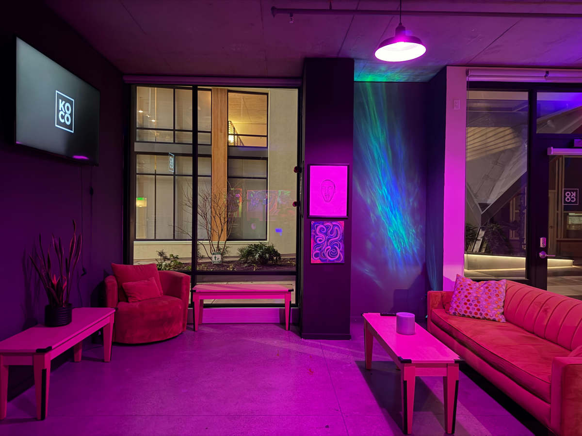 The image shows a modern lounge area with colorful lighting, featuring a red sofa, a light-colored bench, and a decorated wall with artwork.