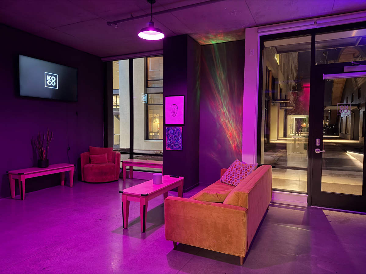 The image shows a modern lounge area with purple lighting, featuring two orange couches, a coffee table, and a large window displaying an outdoor space.
