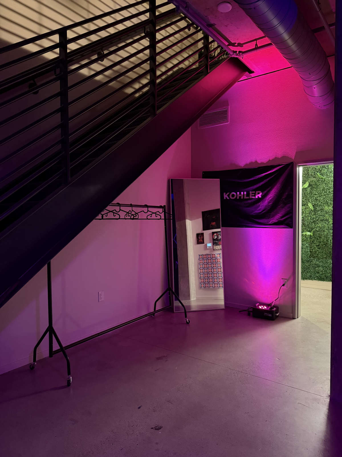 The image shows a modern interior space with a staircase, a full-length mirror, a clothing rack on wheels, and a wall featuring Kohler branding illuminated by purple lights.