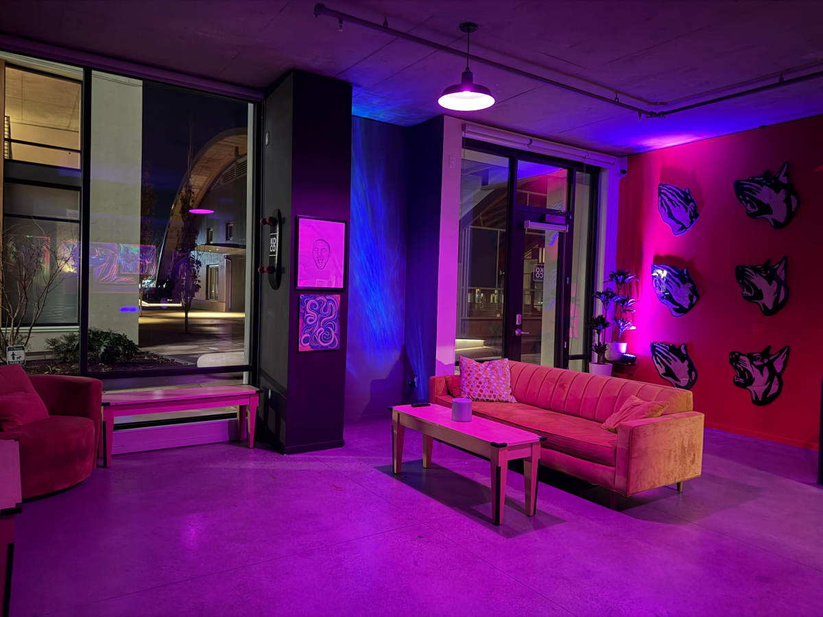 The image shows a modern interior lounge area with a pink couch, a wooden coffee table, and colorful lighting, featuring large windows that overlook an urban setting at night.
