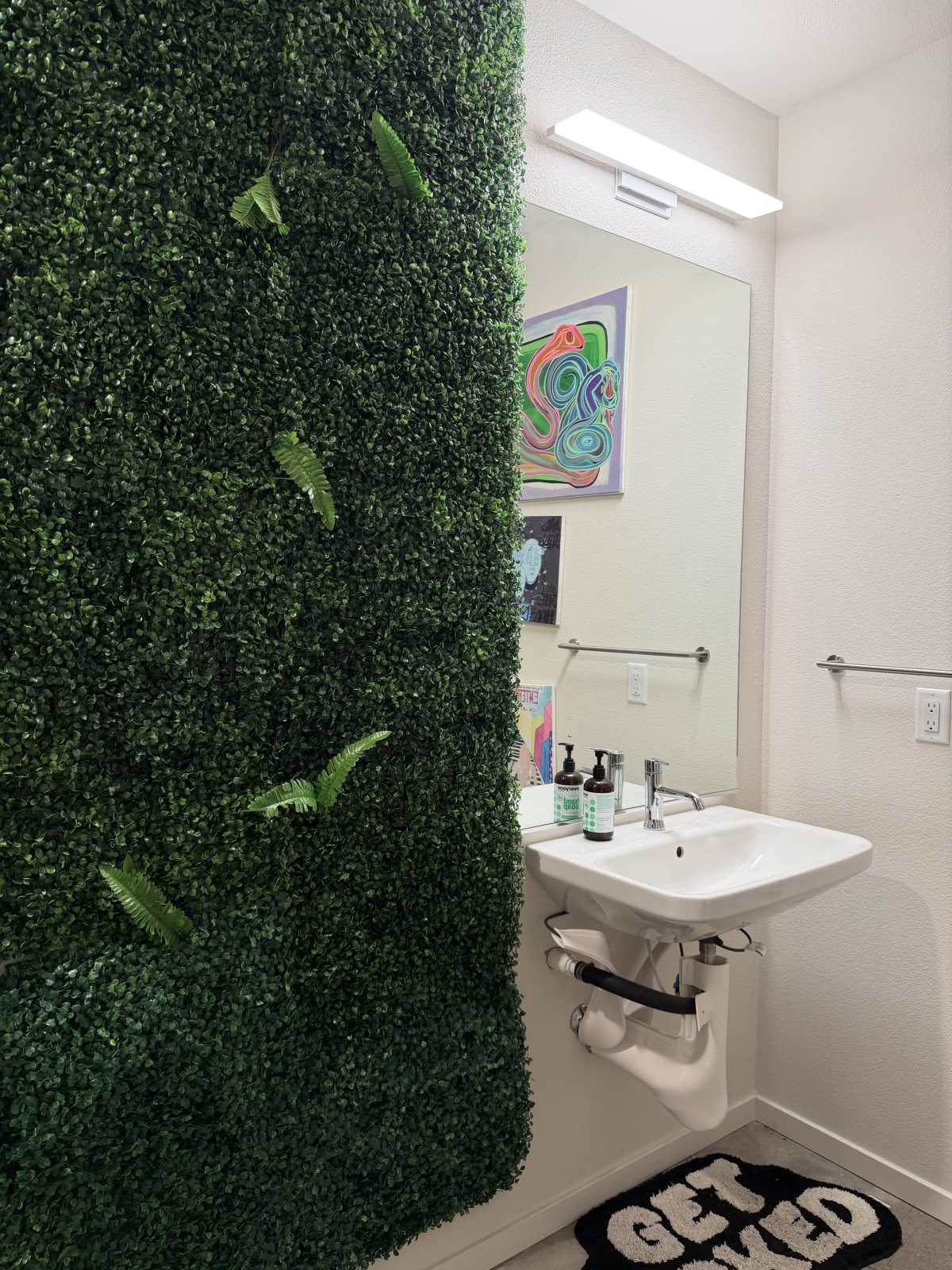 The image shows a modern bathroom with a green wall featuring artificial foliage, a sink with toiletries, and a mirror above it.
