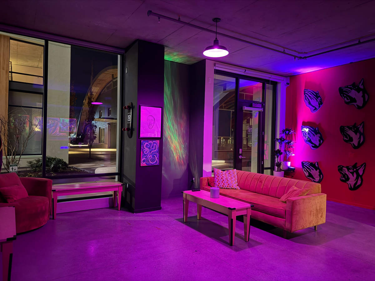 The image shows a lounge area with a pink couch and a coffee table, illuminated by colorful lights reflecting on the walls and a large window revealing a nighttime outdoor scene.