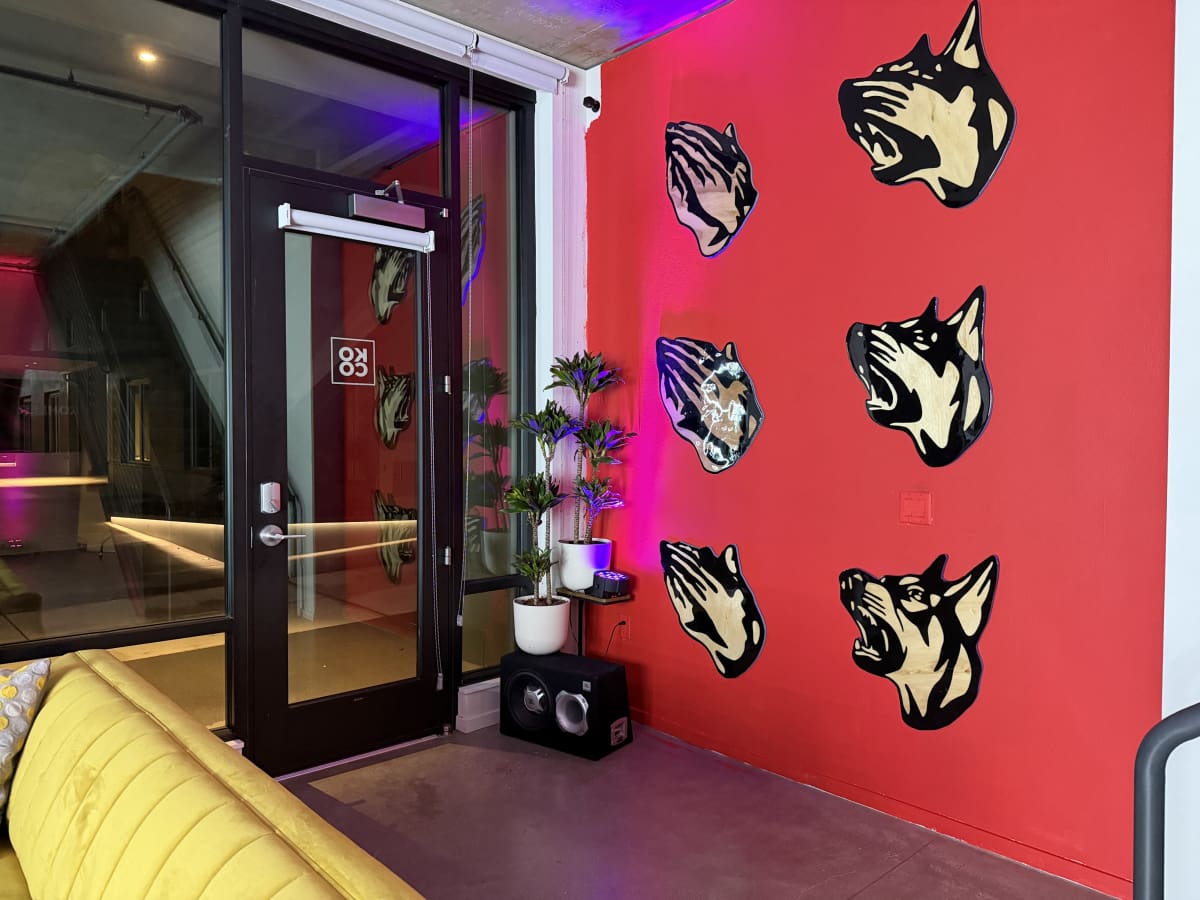 The image shows a modern indoor space with a red wall featuring stylized dog head wall art, a potted plant, and a yellow couch in the foreground.