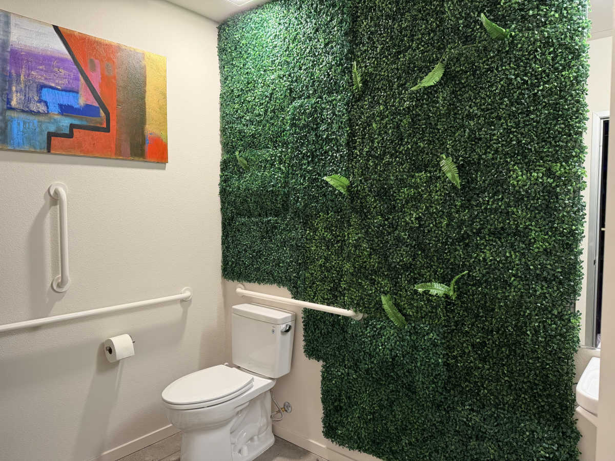 The image shows a bathroom with a toilet next to a wall covered in artificial greenery and a colorful abstract painting above it.