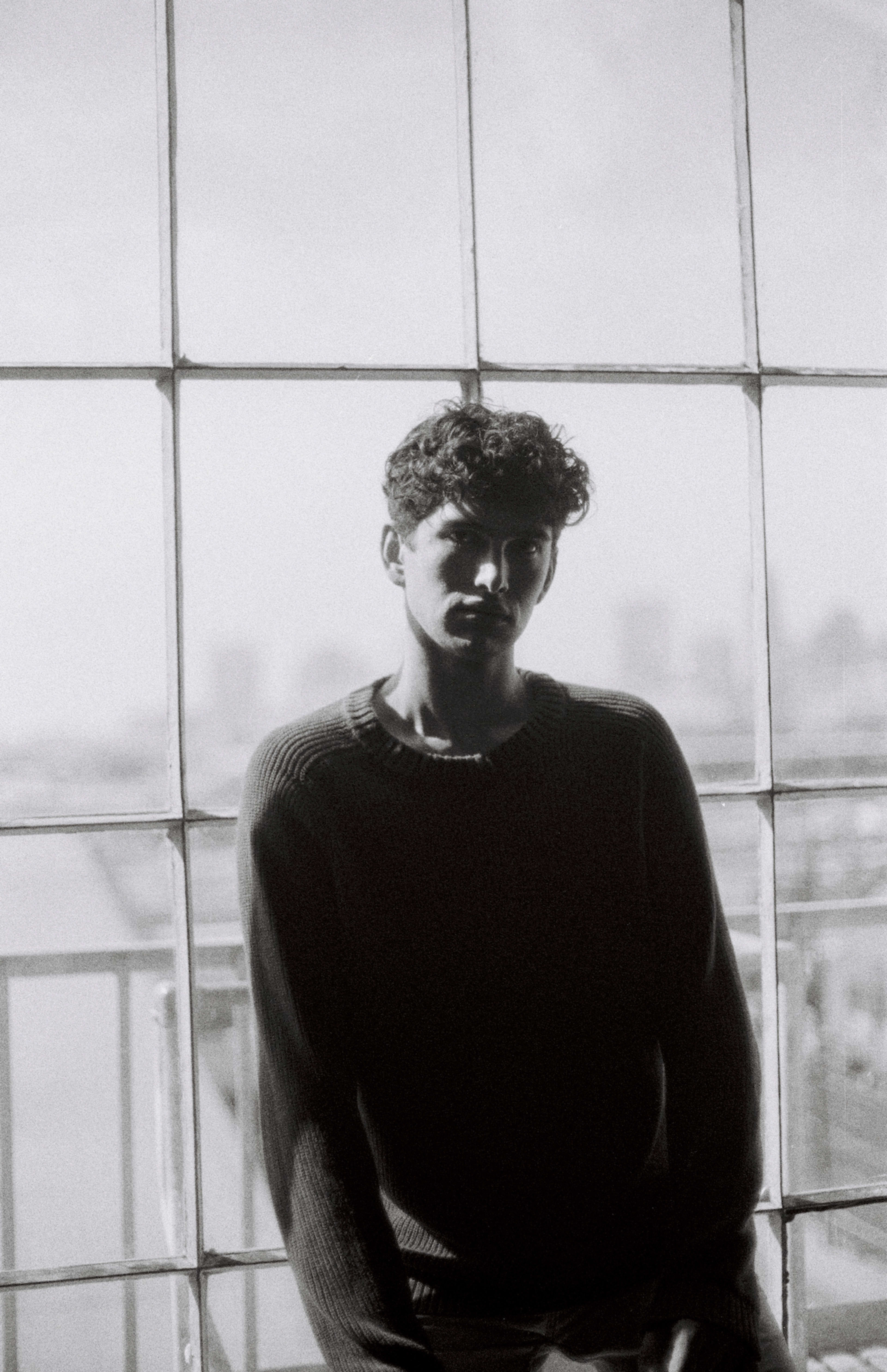 A fashion photoshoot of a man in black and white sitting in front of a window.