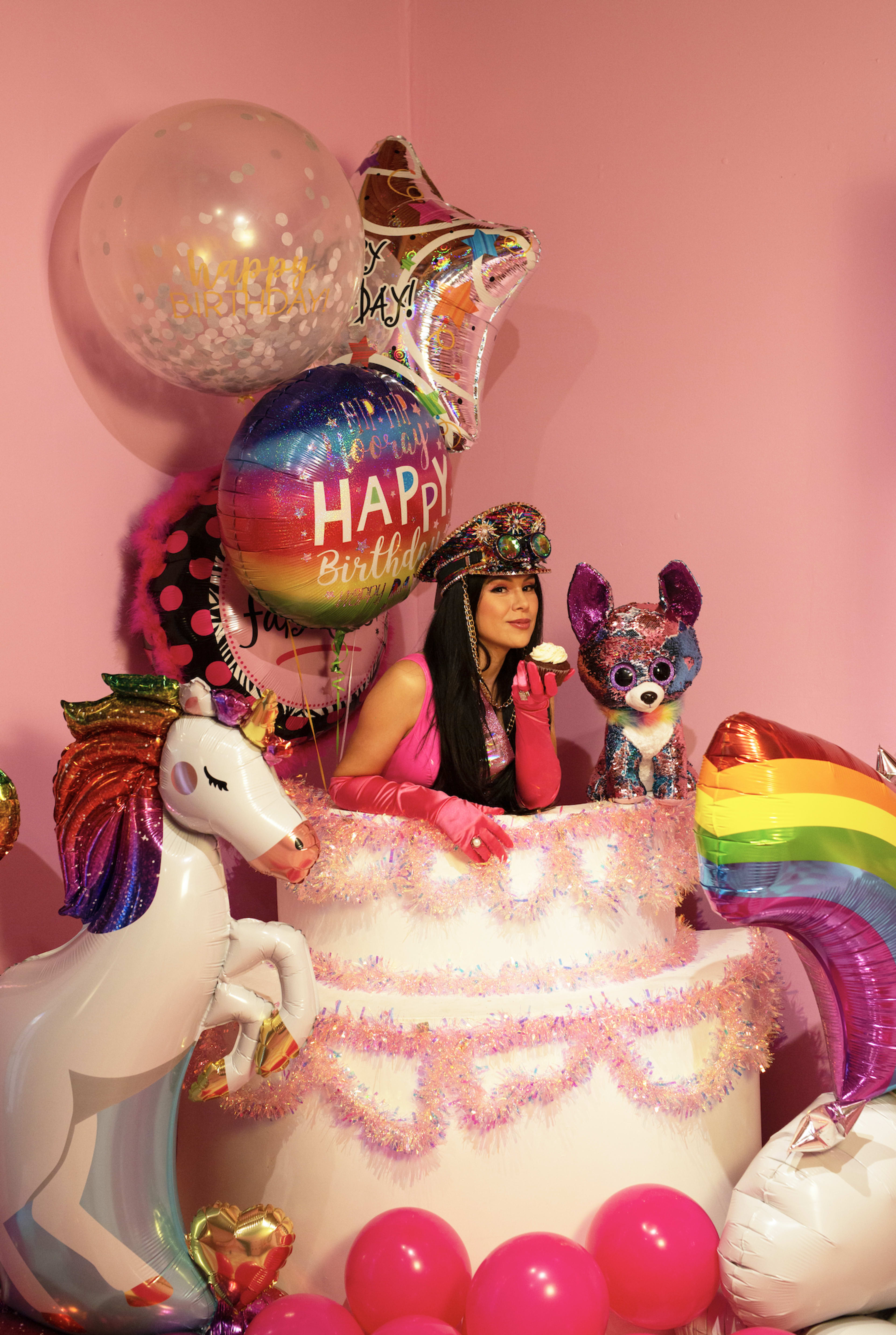 A unicorn-themed birthday photoshoot with a woman sitting on top of a cake surrounded by balloons.