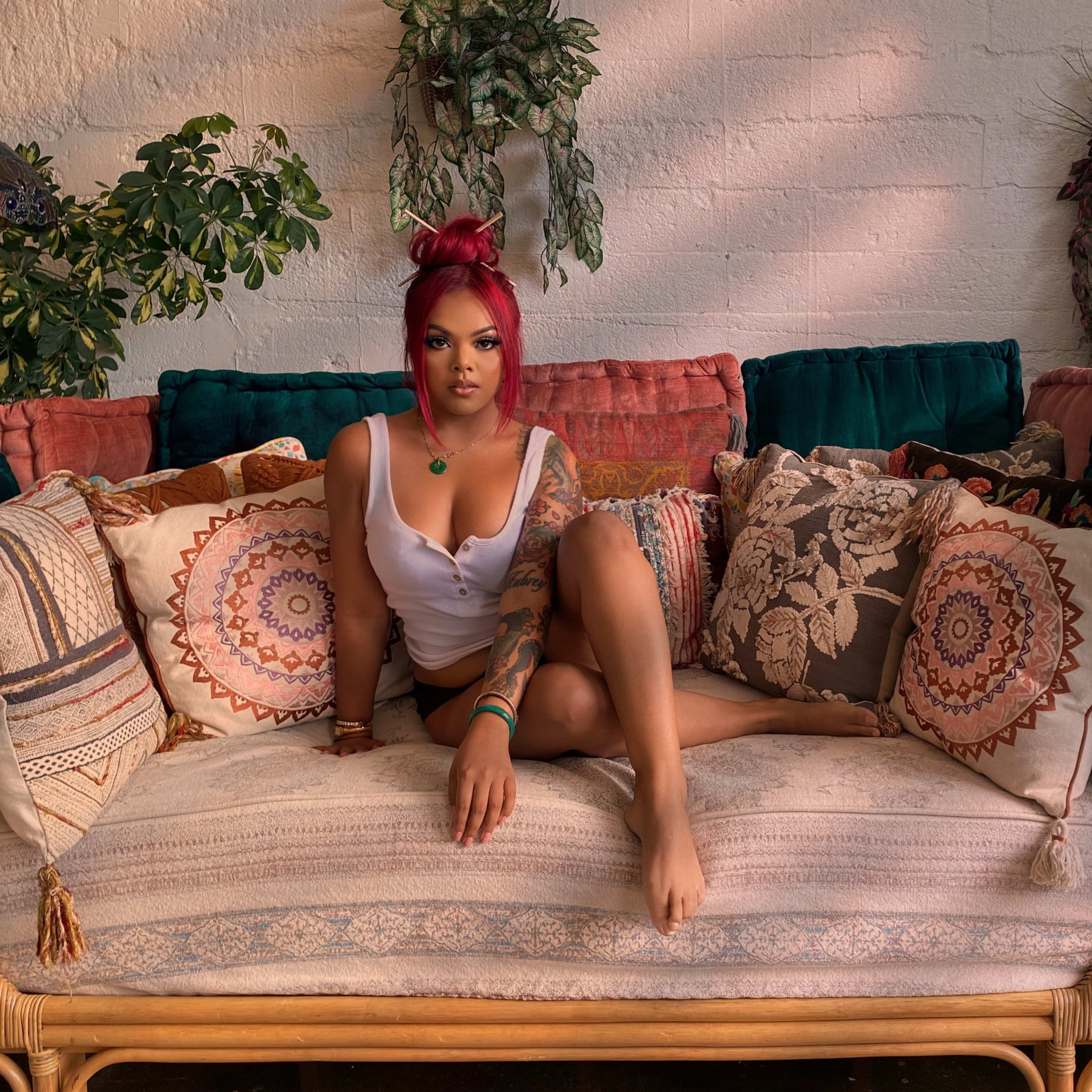 A woman with red hair lounging on a boho couch surrounded by plants during a photo shoot.