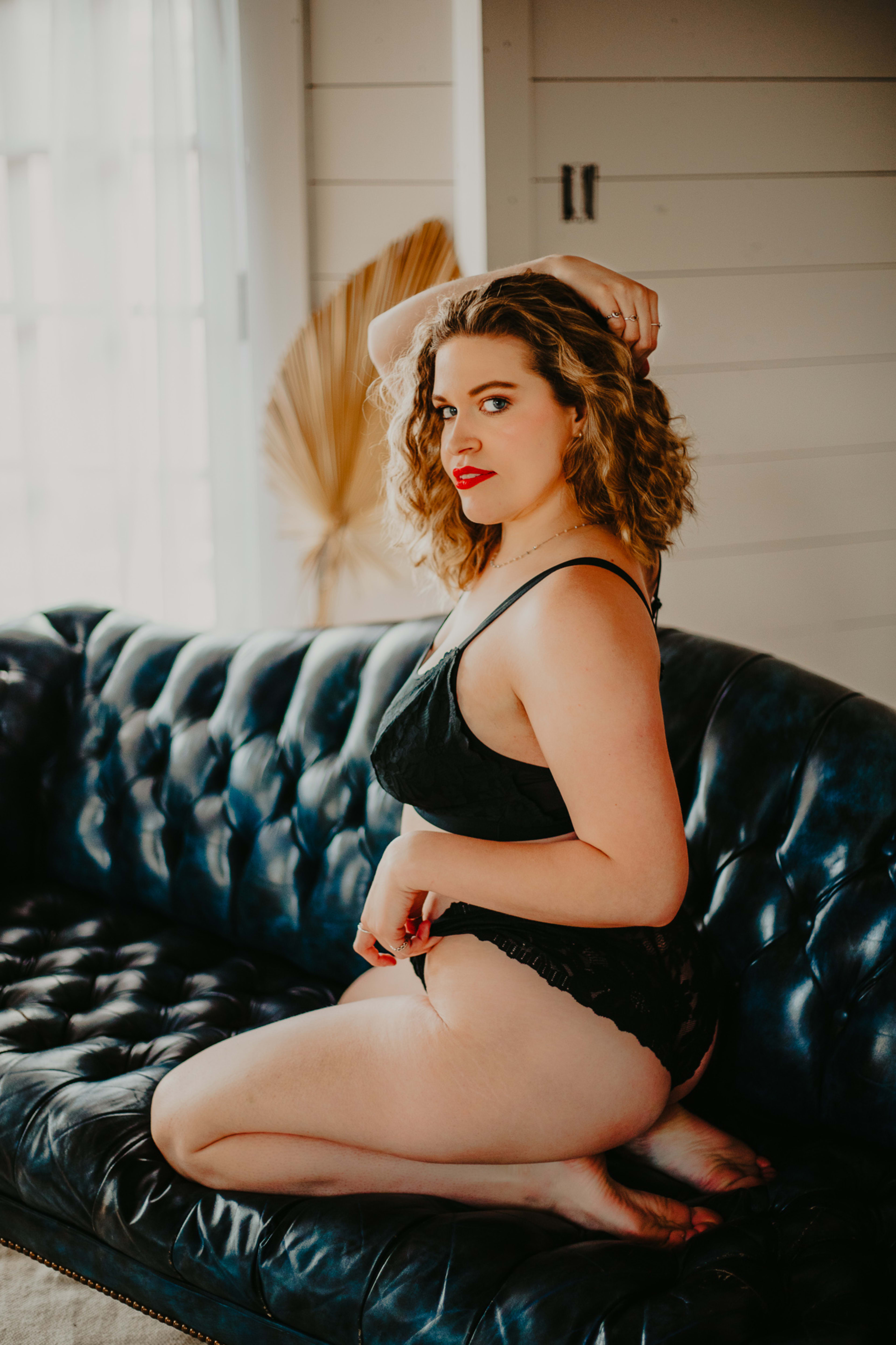 A boudoir photo shoot of a woman in black lingerie sitting on a black leather couch.