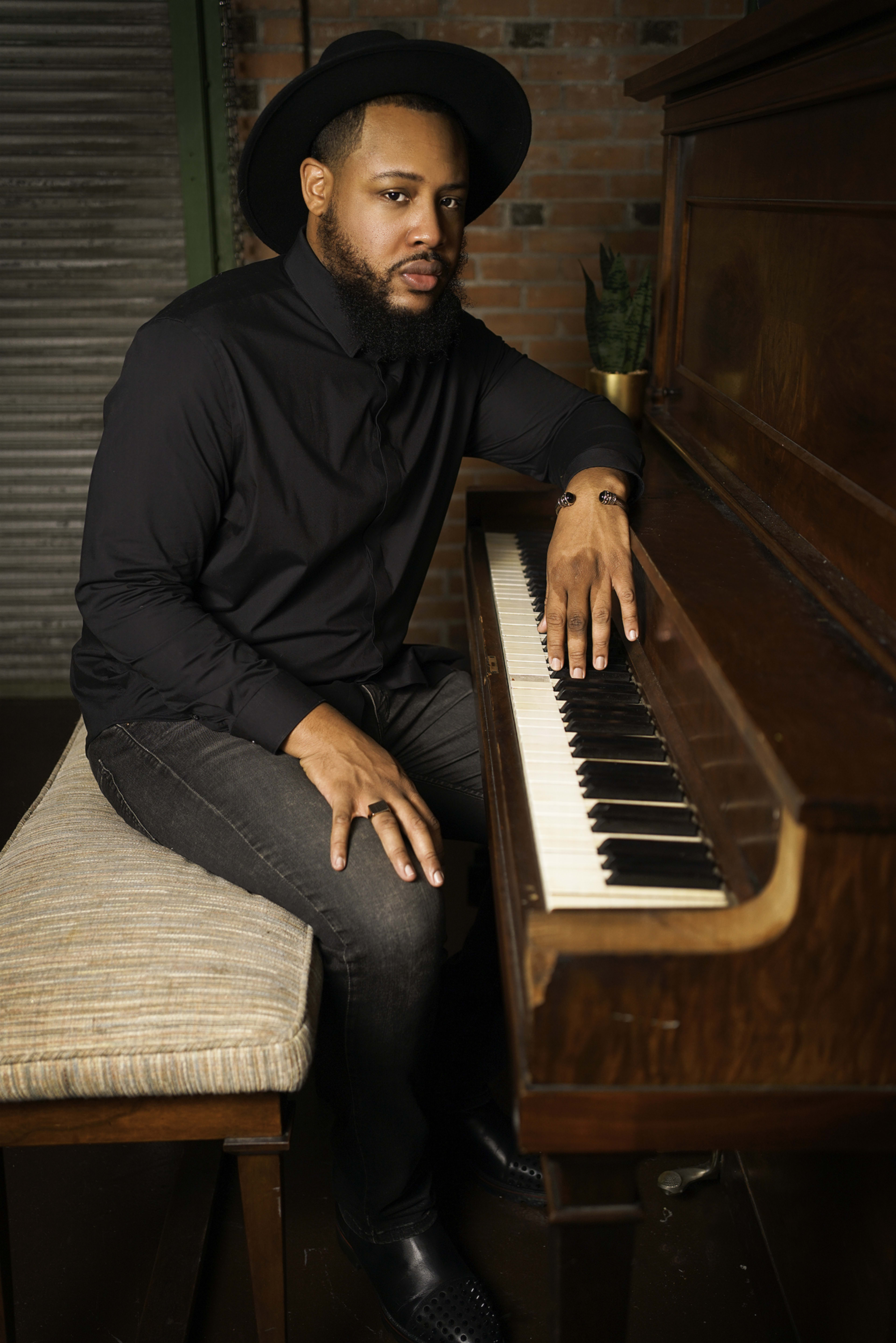 A country musician sitting in front of a brown piano wearing a black hat for his portrait head shot photo shoot.