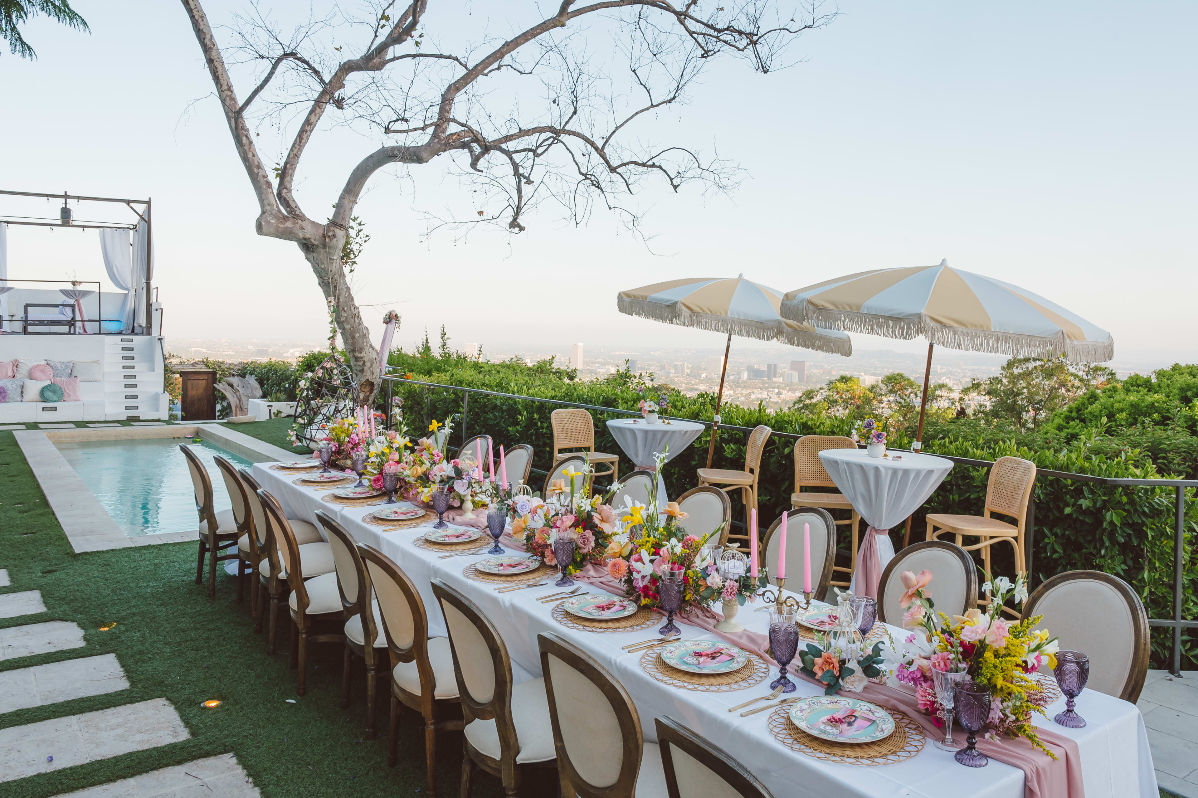 A long table set up for an outdoor party by the pool and overlooking a city view.