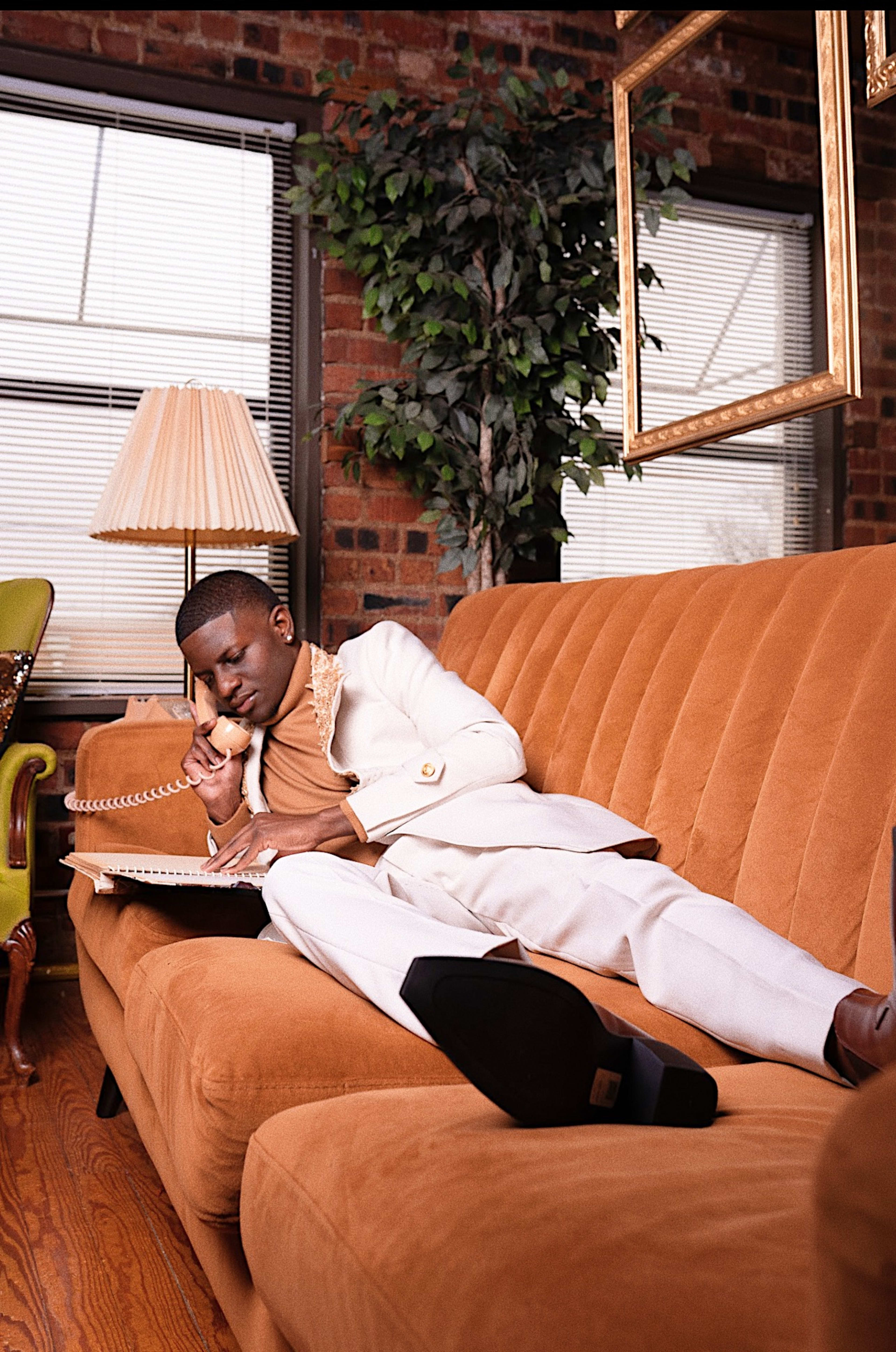A man in a white suit talking on a phone while lying on a retro couch.