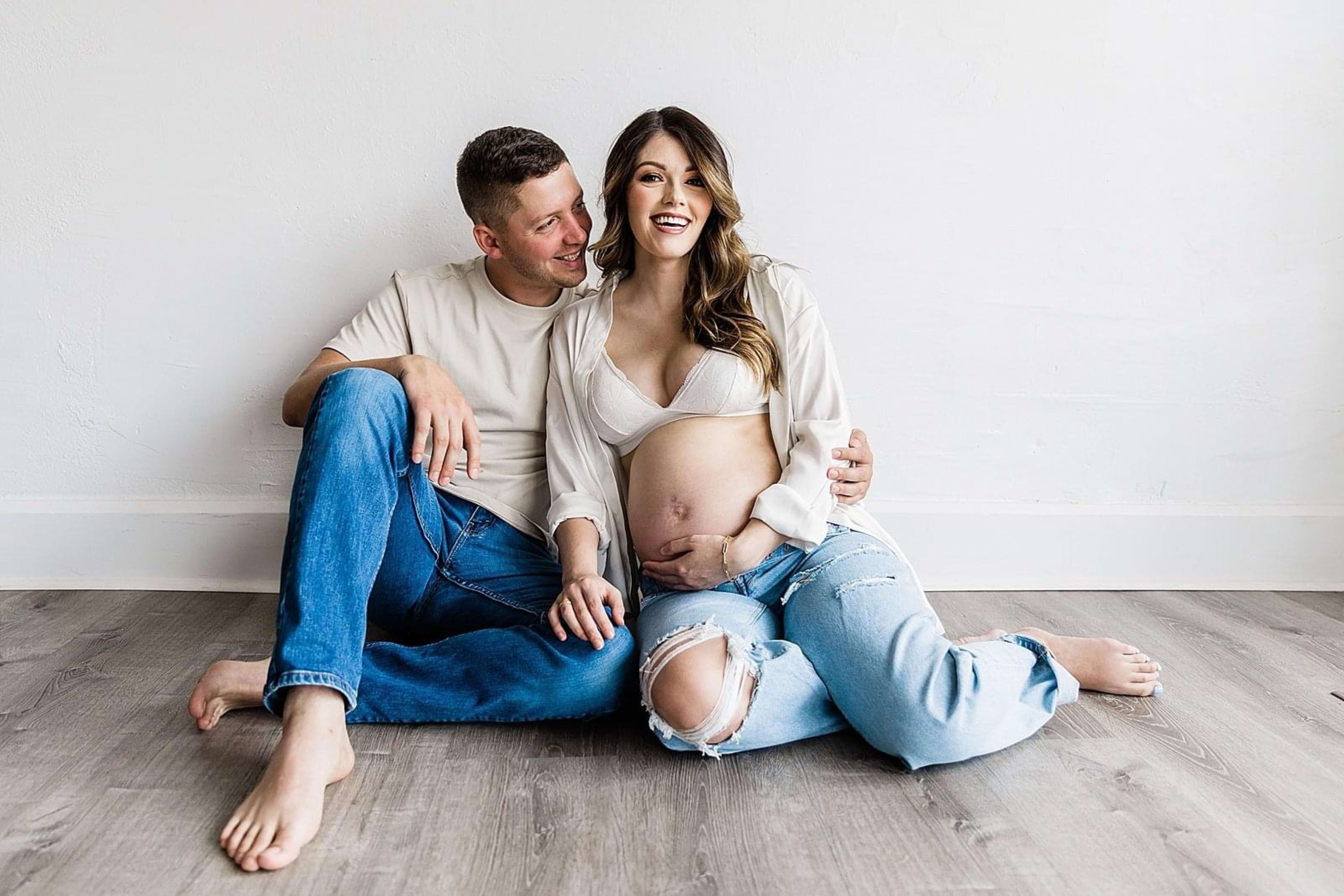 Un shooting photo minimaliste d'un couple attendant un enfant assis sur le sol blanc.