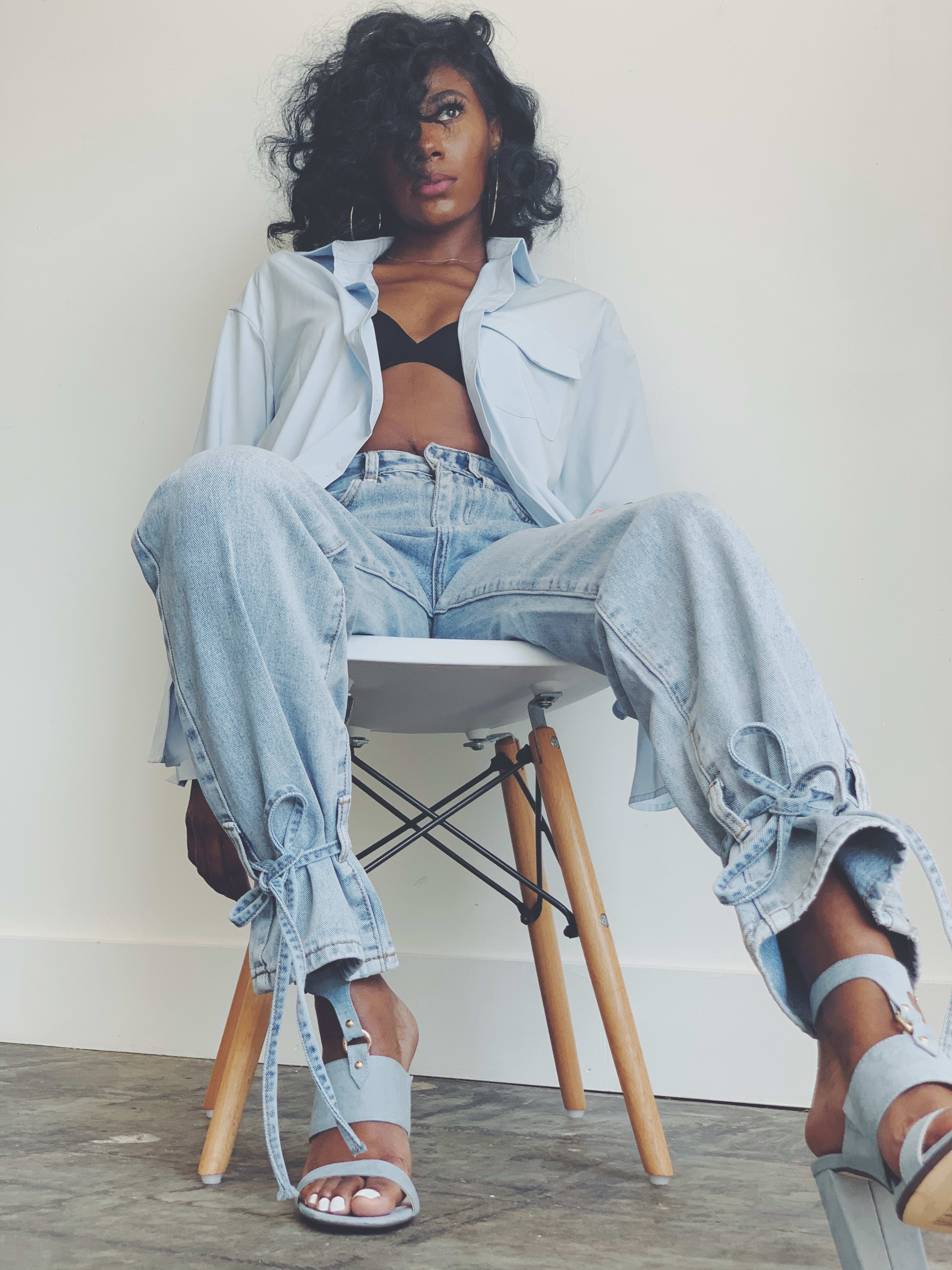 Une femme vêtue d'un jean et d'une chemise bleu clair posant sur une chaise blanche lors d'un shooting photo sur le thème des années 1990.