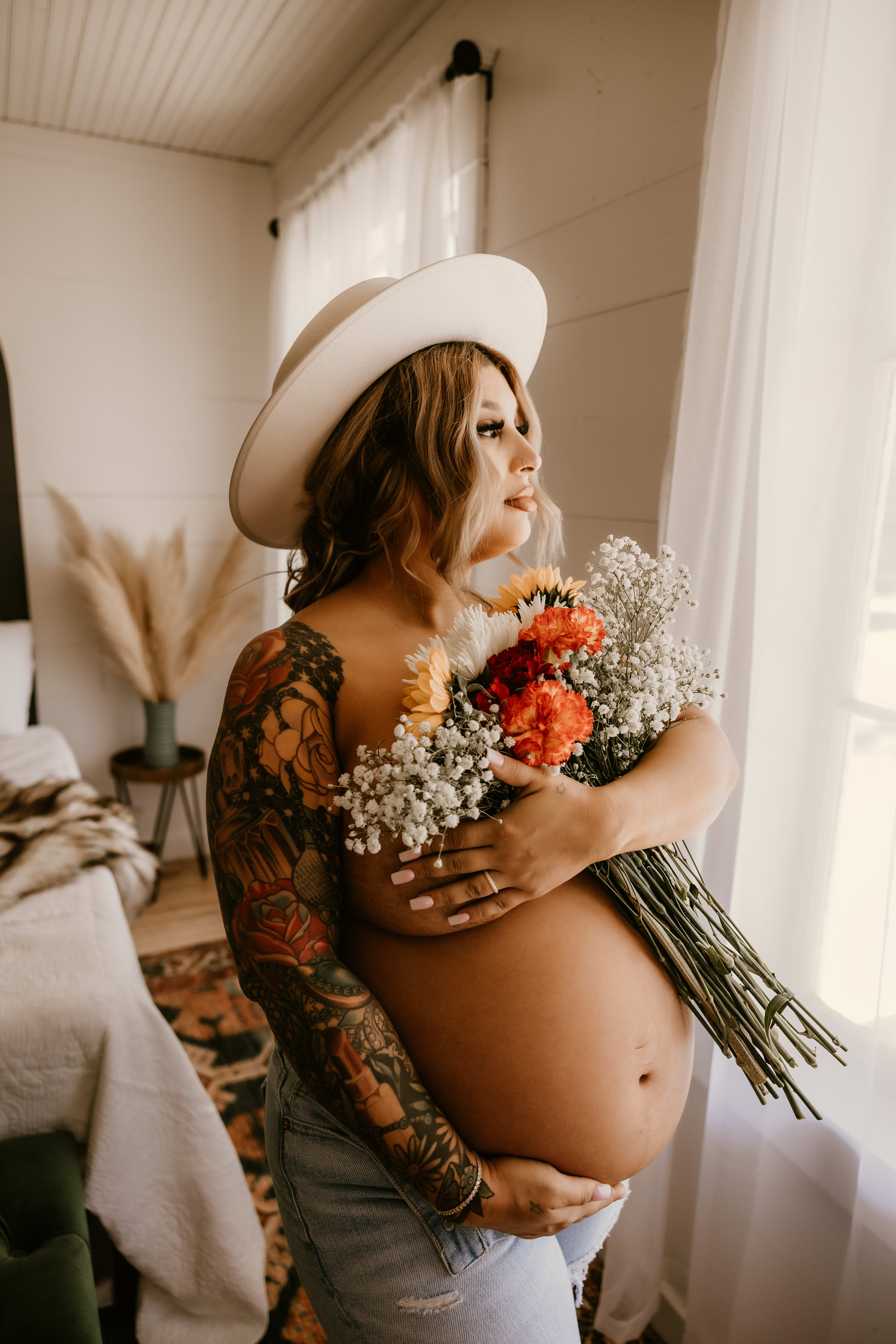 Une femme enceinte portant un chapeau blanc et tenant des fleurs pour son shooting photo de maternité de style boho.