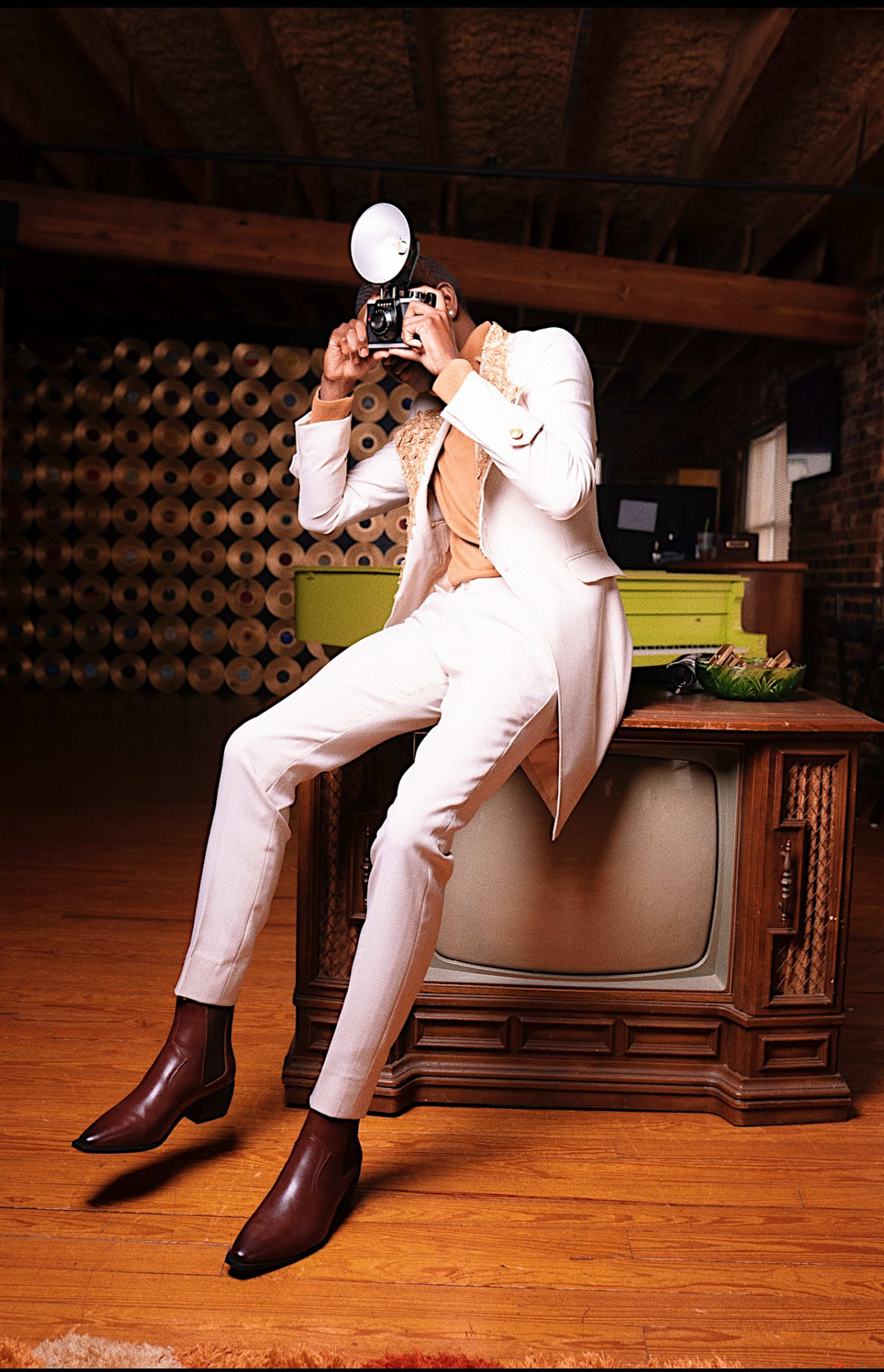 Ein Mann in einem weißen Anzug, der während eines Retro-Fotoshootings auf einem braunen Fernseher posiert.